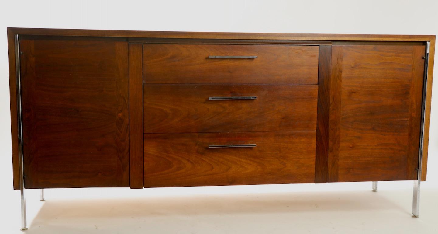 Stylish credenza of walnut and chrome, having two doors flanking a bank of three drawers. This example is in good original condition, clean and ready to use. Design in the style of Milo Baughman, unsigned.
