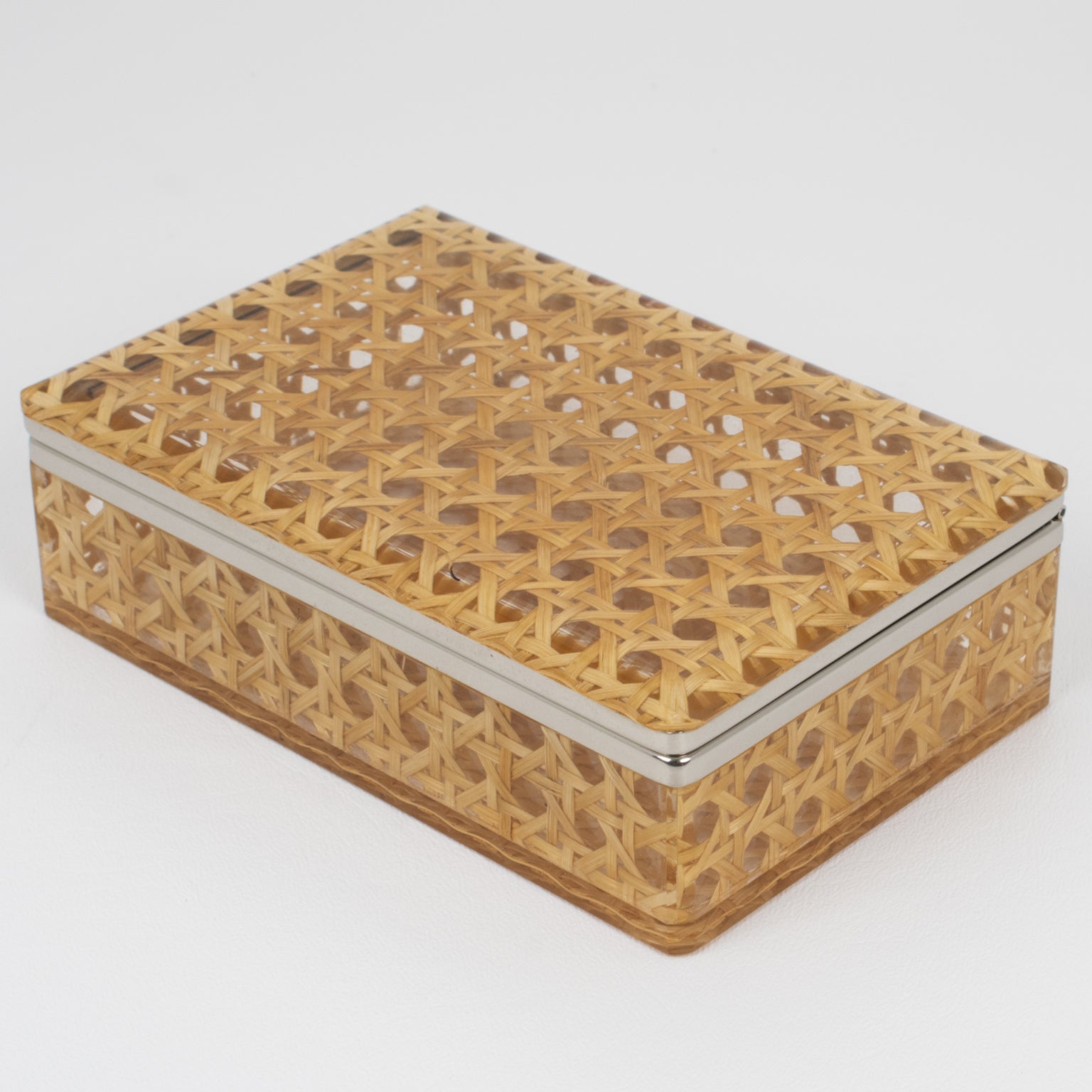 This lovely decorative hinged box is perfect for a living space or vanity. The chrome-plated framing is complemented with clear Lucite and rattan or wicker cane work. The rattan is embedded within two thick sheets of clear Lucite or acrylic material