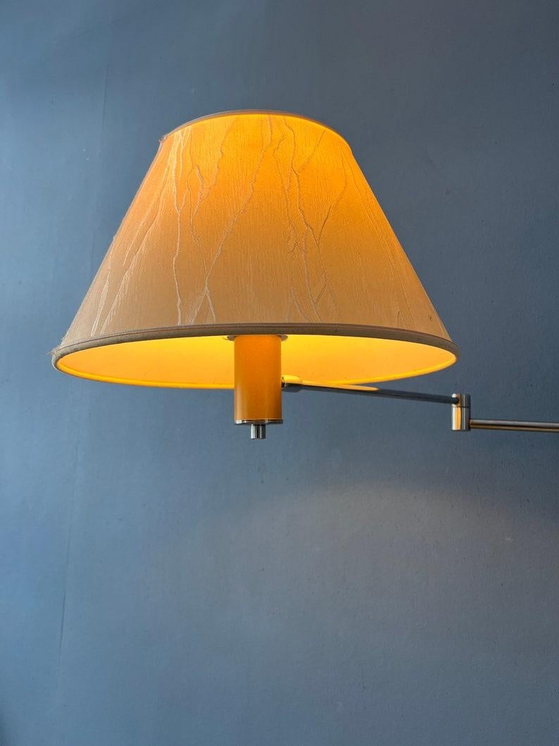 Classic mid-century wall lamp with chrome swing-arm frame and beige shade. The length and position of the lamp can easily be adjusted with the swing mechanism. The lamp requires an E27/26 (standard) lightbulb and currently has an EU-plug (easily