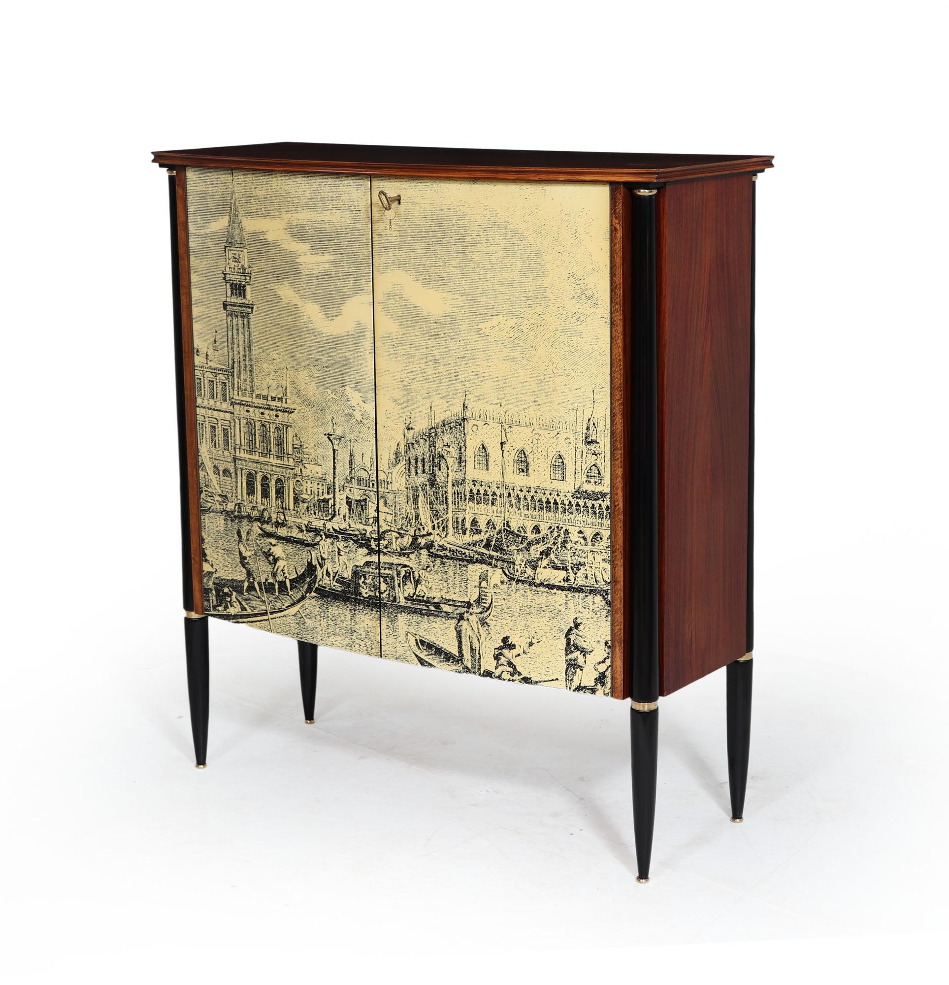 ITALIAN MID CENTURY COCKTAIL CABINET
Produced in Italy in the 1950’s in the Manner of Piero Fornasetti the walnut cabinet is supported by ebonised turned legs with brass detail and feet. To the bowed door front is a lithograph printed classic scene