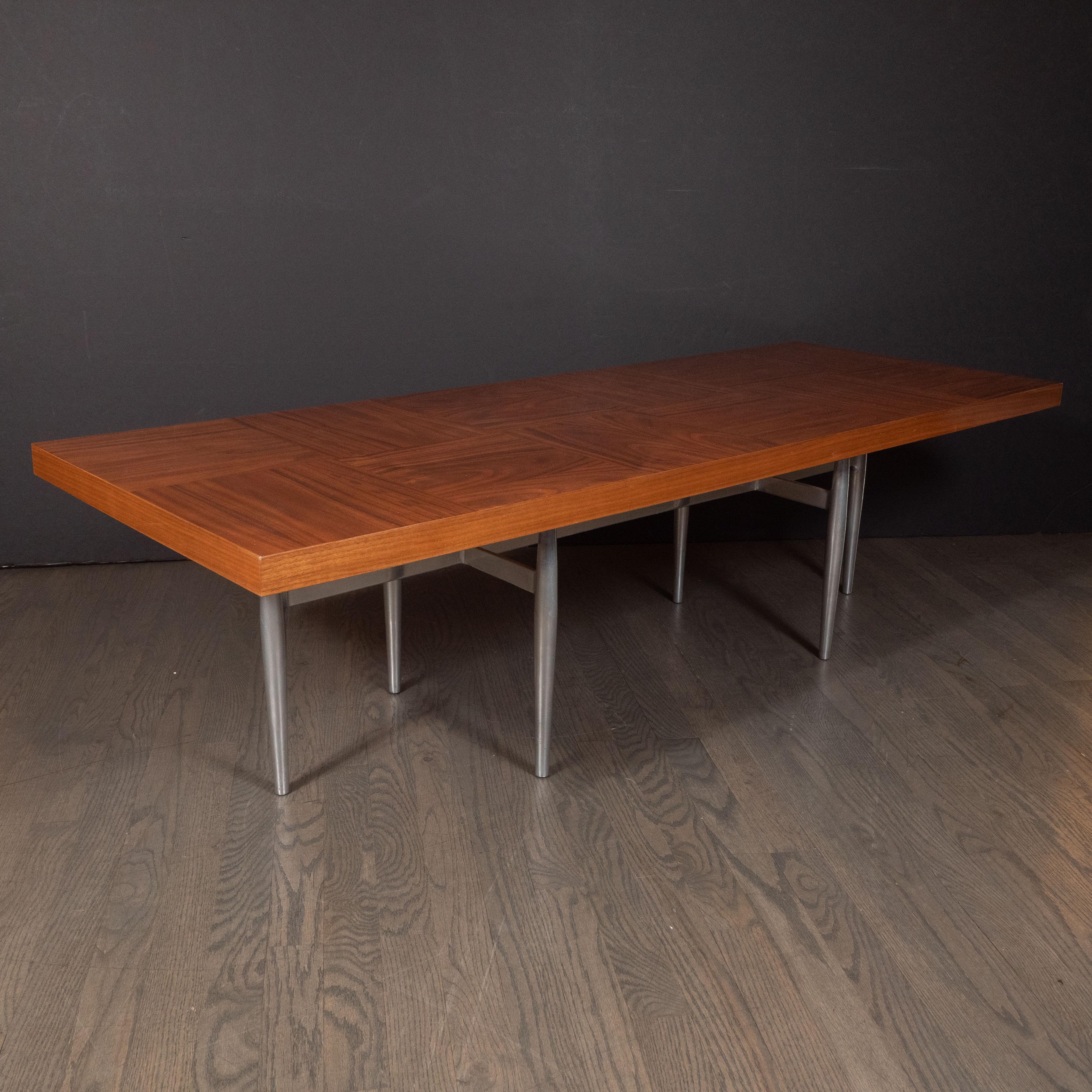 This stunning cocktail table was realized in the United States, circa 1960. It features a sculptural base consisting of six legs with tapered ends connected via rectangular supports all in brushed aluminum. A rectangular bookmatched walnut top- with