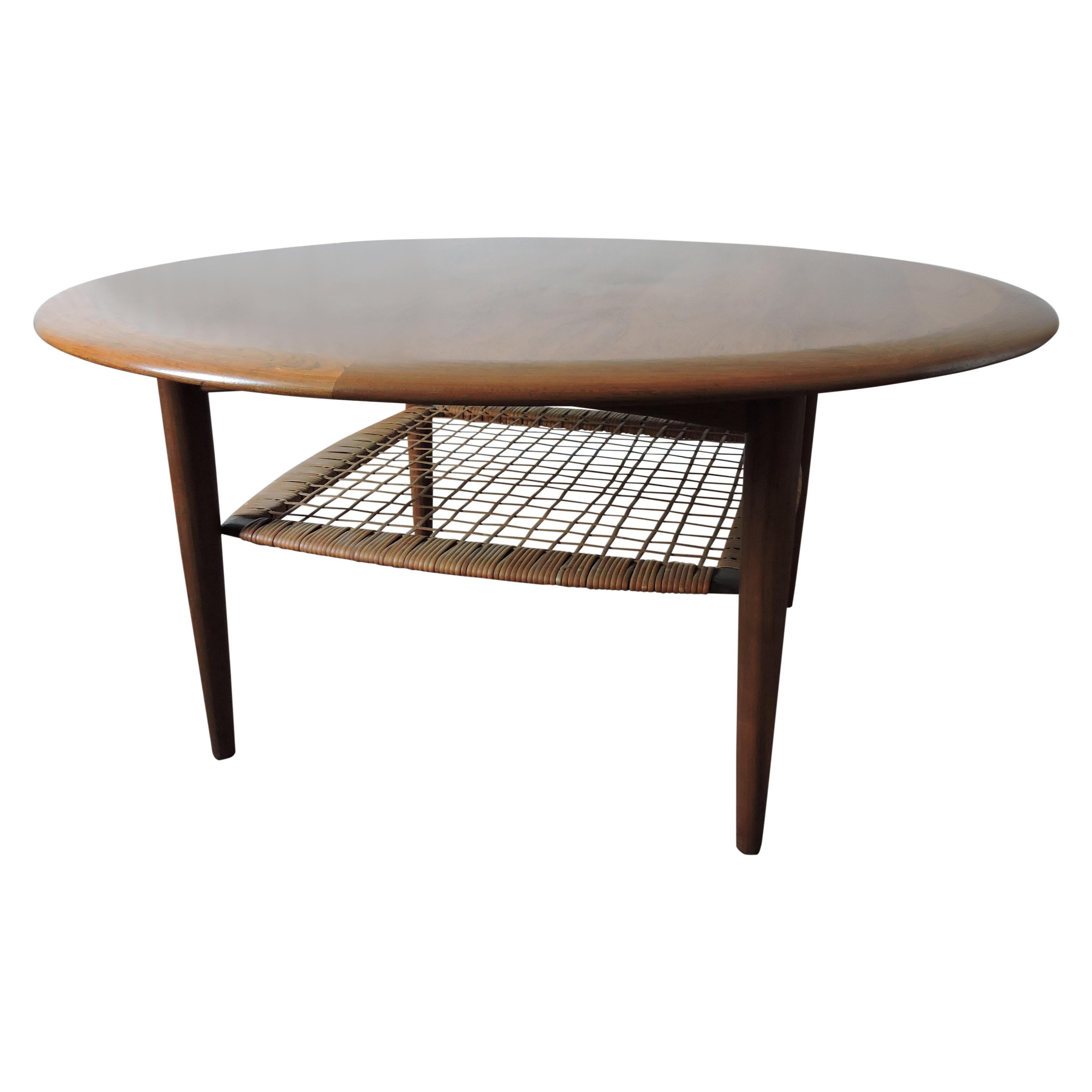 A round coffee table by Johannes Andersen for CFC Silkeborg.
The tabletop is veneered in teak and framed by a wide solid teak edge.
It features a magazine rack made from a solid teak frame covered with woven rattan shelf.
       