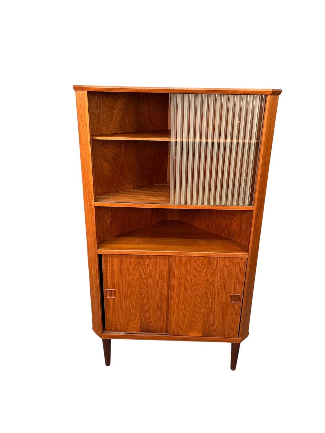 This stunning mid-century teak and glass corner cabinet is sure to be a standout piece in any home. Crafted from high-quality teak wood, this piece is both sturdy and beautiful. The glass doors allow for easy display of your favorite items, while