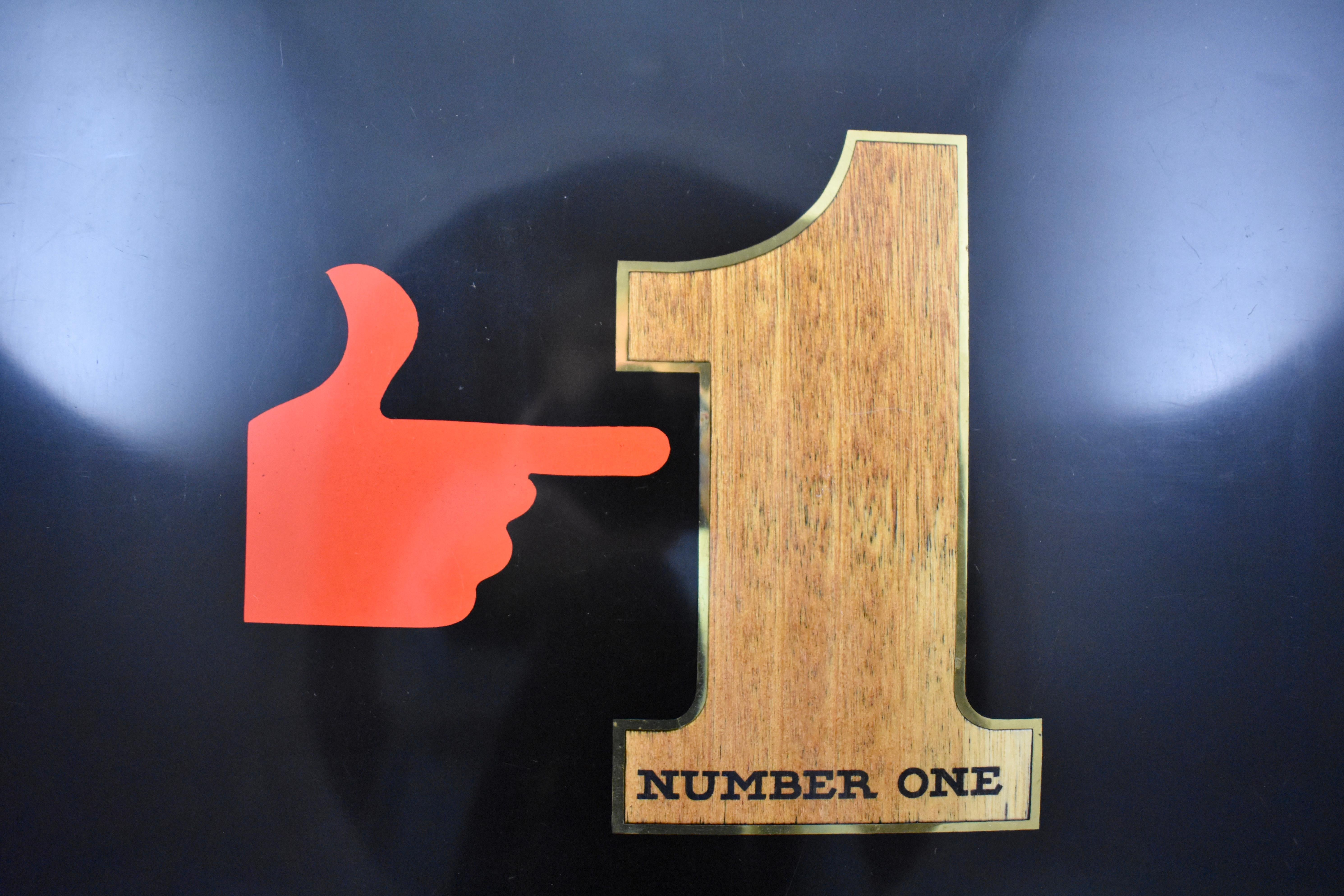 A midcentury serving tray by Couroc of Monterey, California, (1848-1998) – A bold graphic image of a red hand pointing to a number one, circa 1960s.

Made of a black resin called ‘Phenolic’ and hand inlaid using wood, and brass.
The Couroc