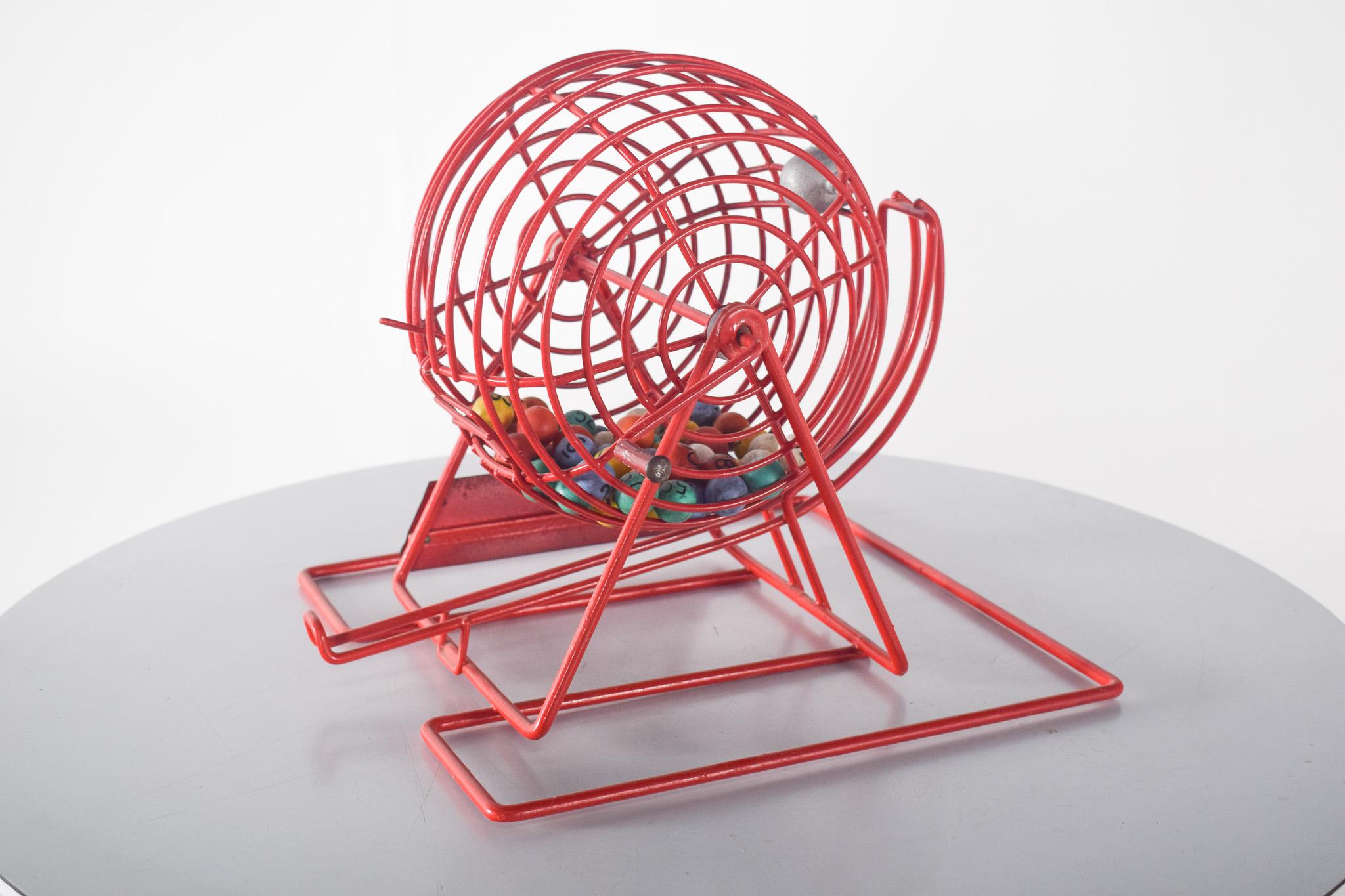 Roulette à manivelle pour le jeu de Bingo. En métal peint en rouge, avec une structure sphérique en forme de cage reposant sur une base solide. Il s'agit d'un accessoire traditionnel utilisé dans les jeux de bingo, dans lequel des boules numérotées