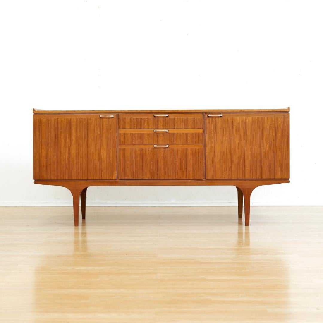 Cette crédence du milieu du siècle a été produite par Meredew à Letchworth dans le Hertfordshire en Angleterre. Fabriqué dans les années 1960, avec trois tiroirs centraux et deux armoires de rangement, il est parfait pour un salon ou une salle à