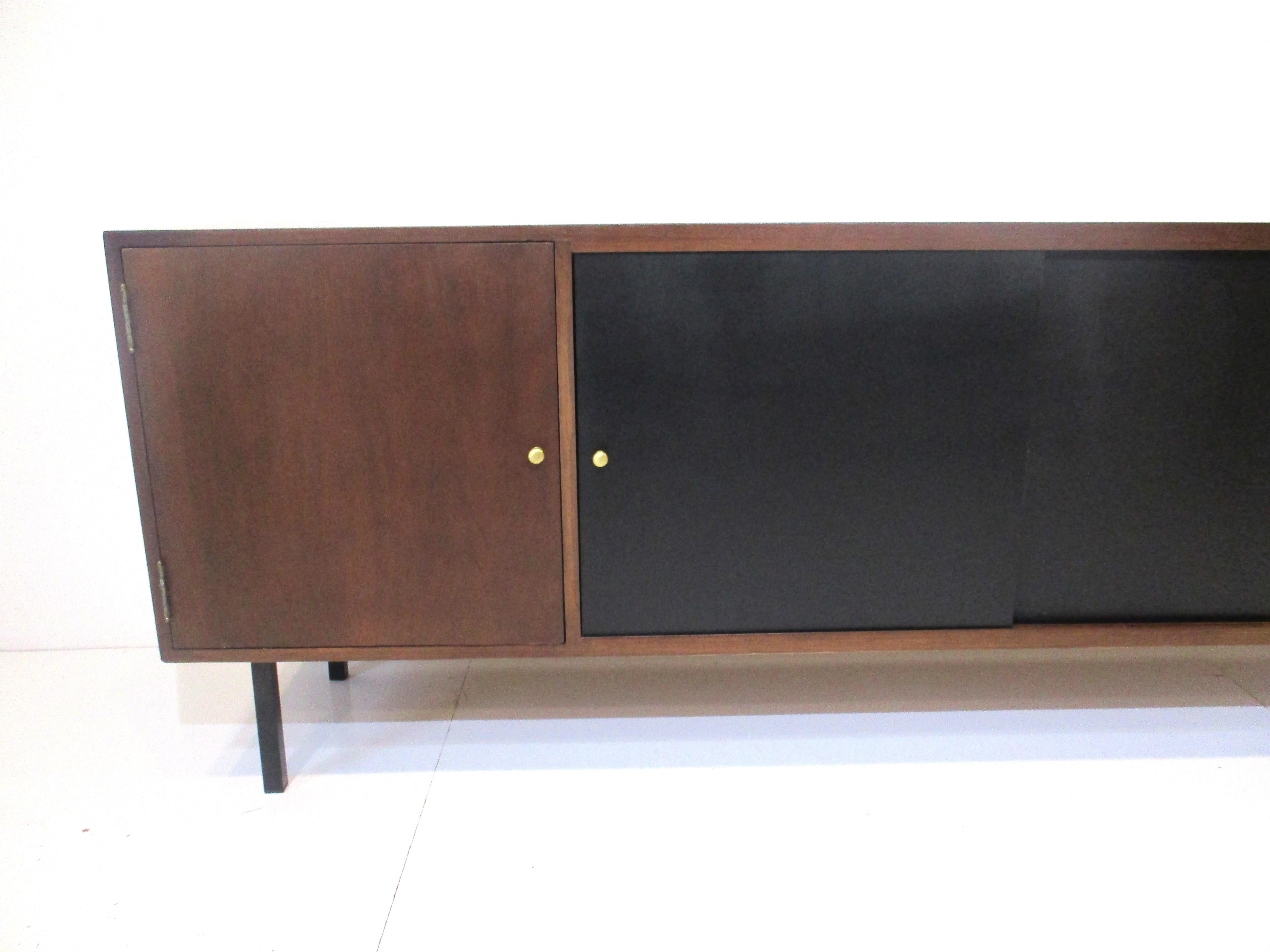 A walnut credenza with two sliding center doors in a satin black finish with brass pulls and two outer doors that swing open. Inside the end storage areas are fixed shelves for storage as is the center shelve all sitting on black steel legs designed