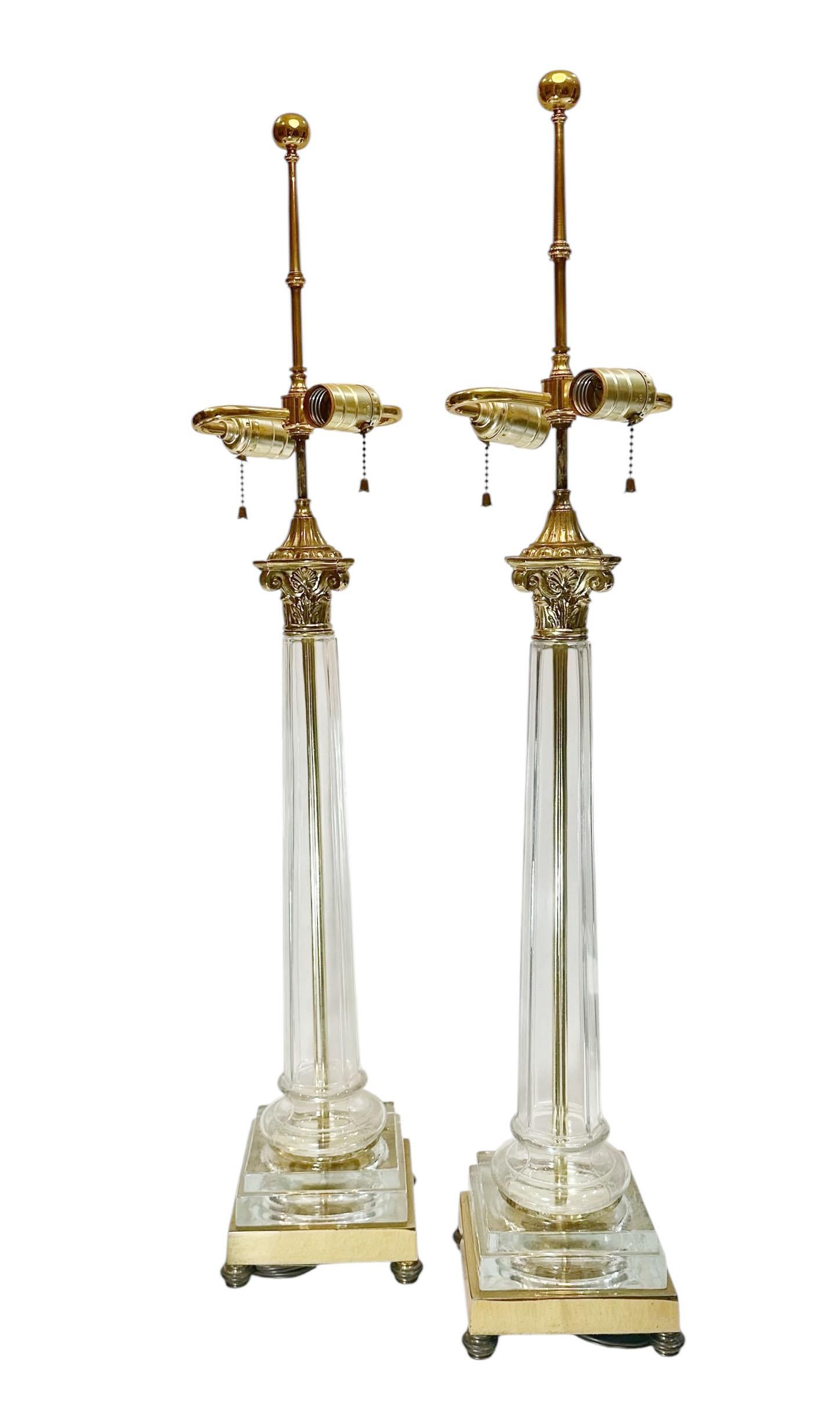 Un llamativo par de lámparas de cristal y latón de mediados del siglo XX, diseñadas con inspiración clásica. Cada lámpara está fabricada con un cuerpo de cristal transparente acentuado con detalles de latón, que adopta la forma de una columna que