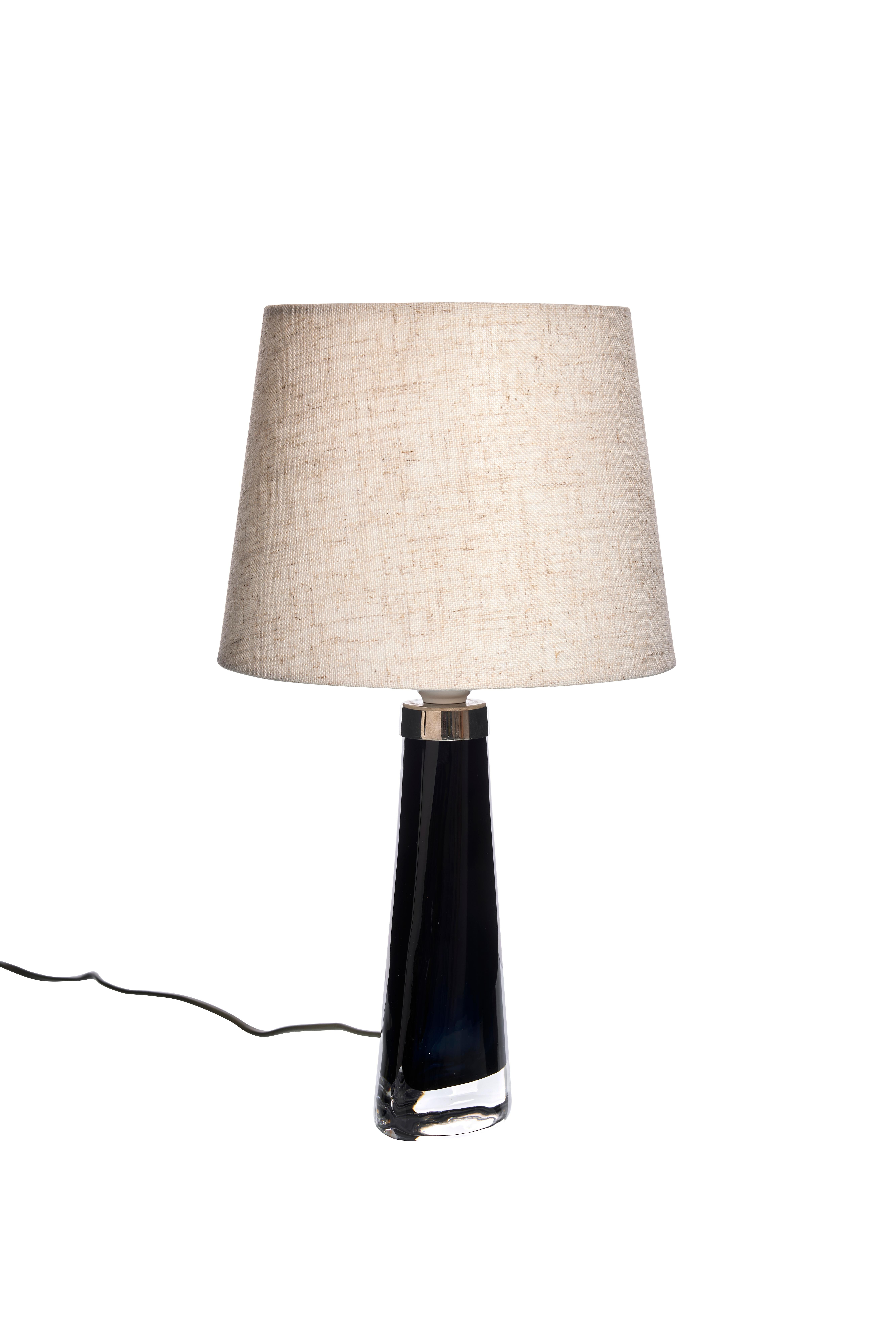 Esta lámpara de sobremesa presenta una base de cristal elegante y cónica en un tono azul intenso con una superficie pulida que añade elegancia a cualquier espacio. El cristal pasa suavemente a un acabado transparente en la parte inferior,