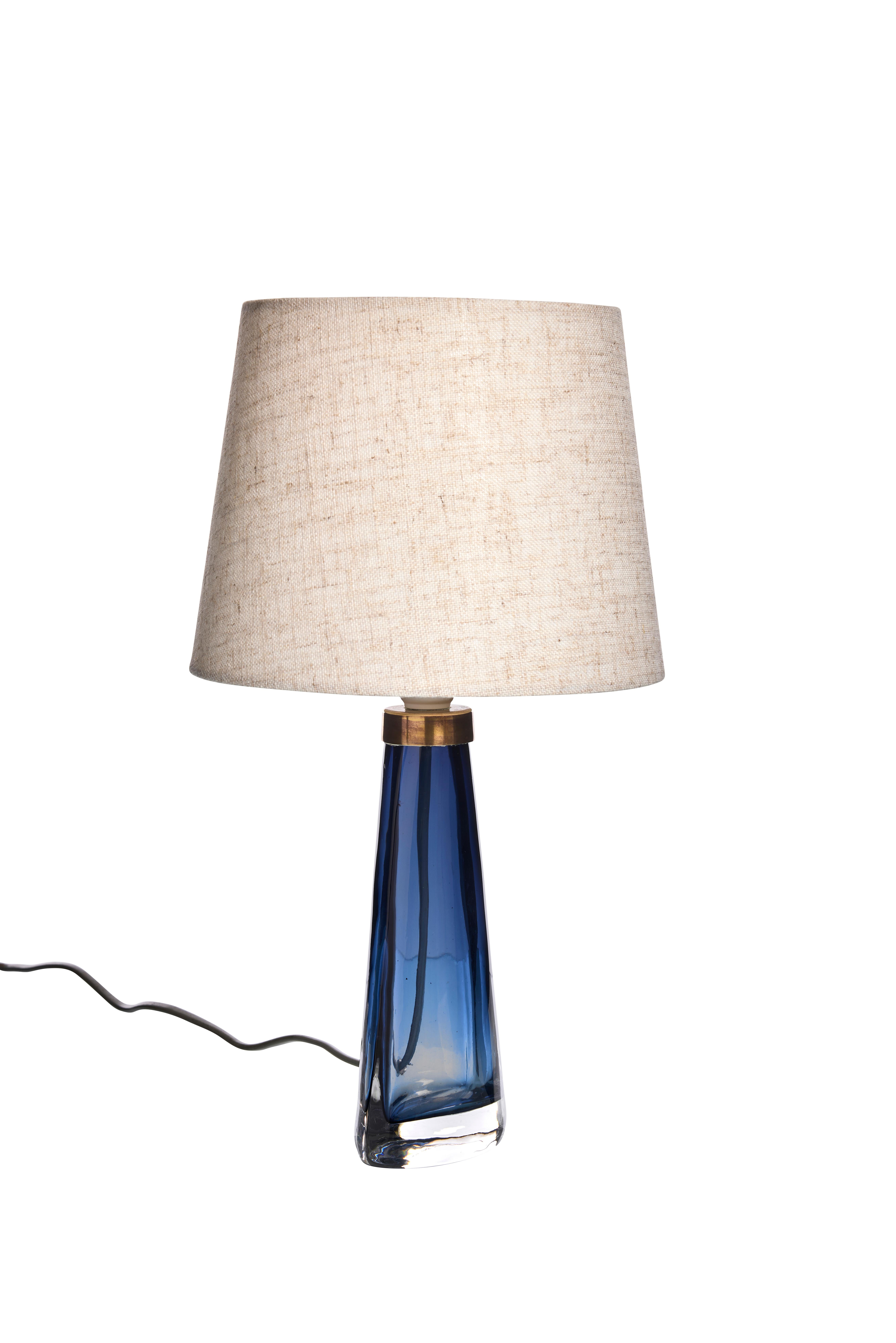 Esta impresionante lámpara de sobremesa presenta una base de cristal azul bellamente elaborada con una tapa de latón. Su silueta elegante y afilada añade un toque de elegancia a cualquier espacio. Ten en cuenta que la pantalla de la lámpara no está