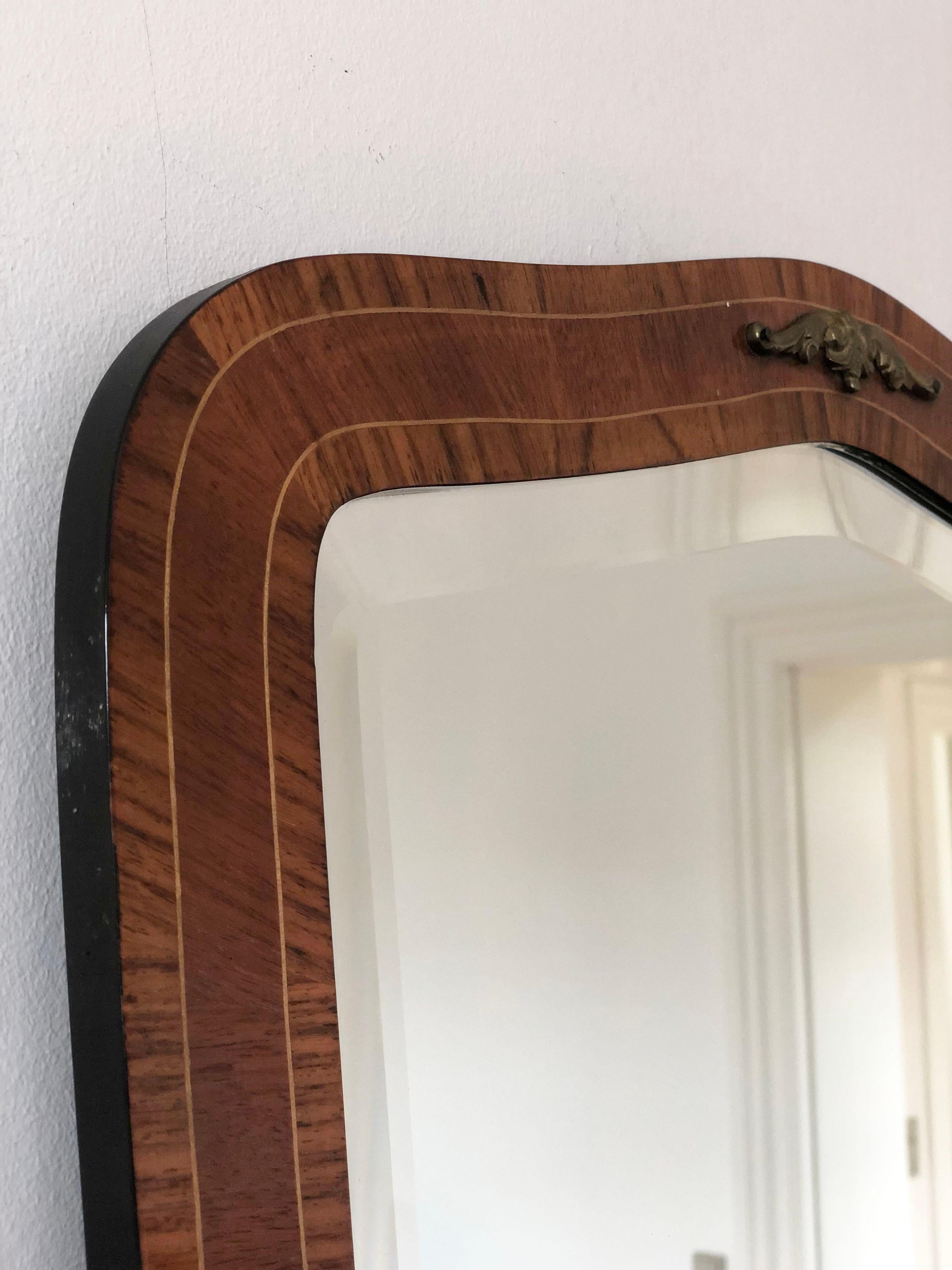 Mid-century wall mirror from Denmark with curved wooden frame and bevelled glass. The frame is made of mahogany veneer with maple inlay lines and has a decorative brass ornament at the top. 

Object: mirror
Style: Vintage
Period: 1950-1960
Country