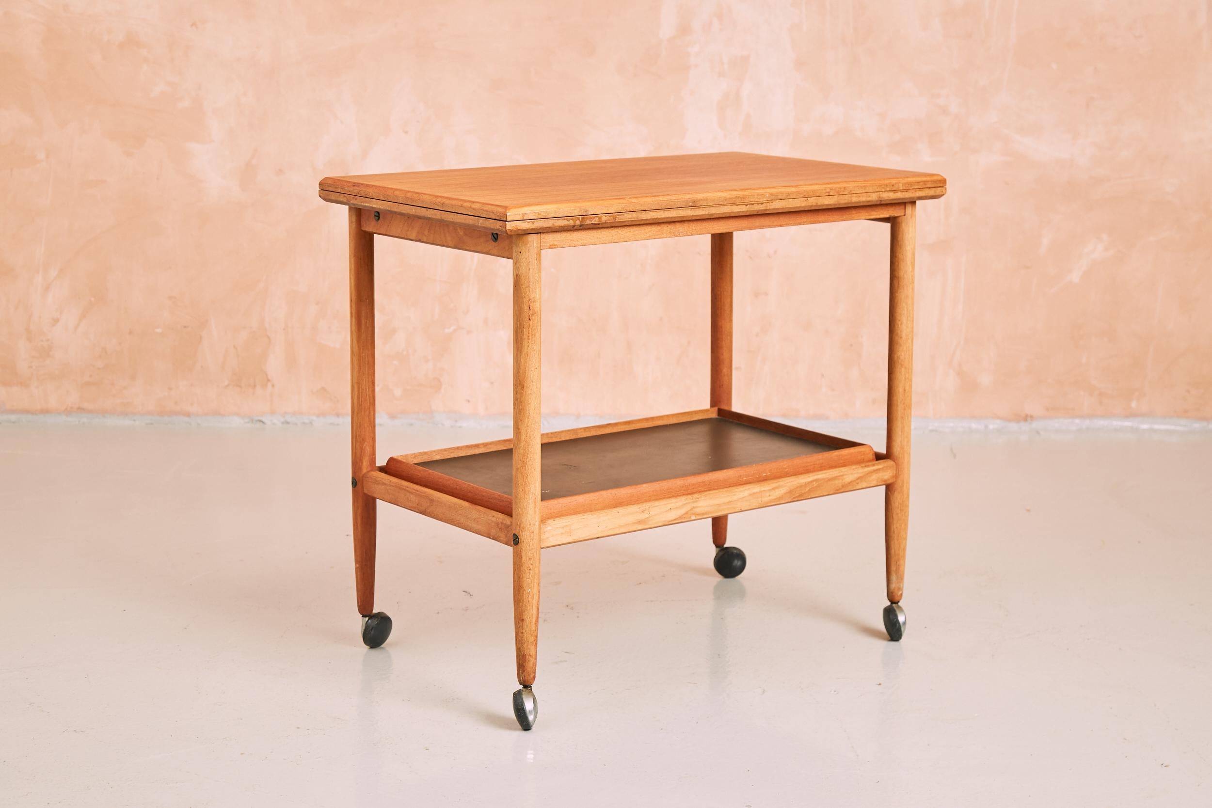 A vintage Danish side table/serving trolley set on casters with fold-out top and removable serving tray. This convenient and rare piece of mid century design is produced in teak and features a revolving, fold-out top, with attractive exposed brass