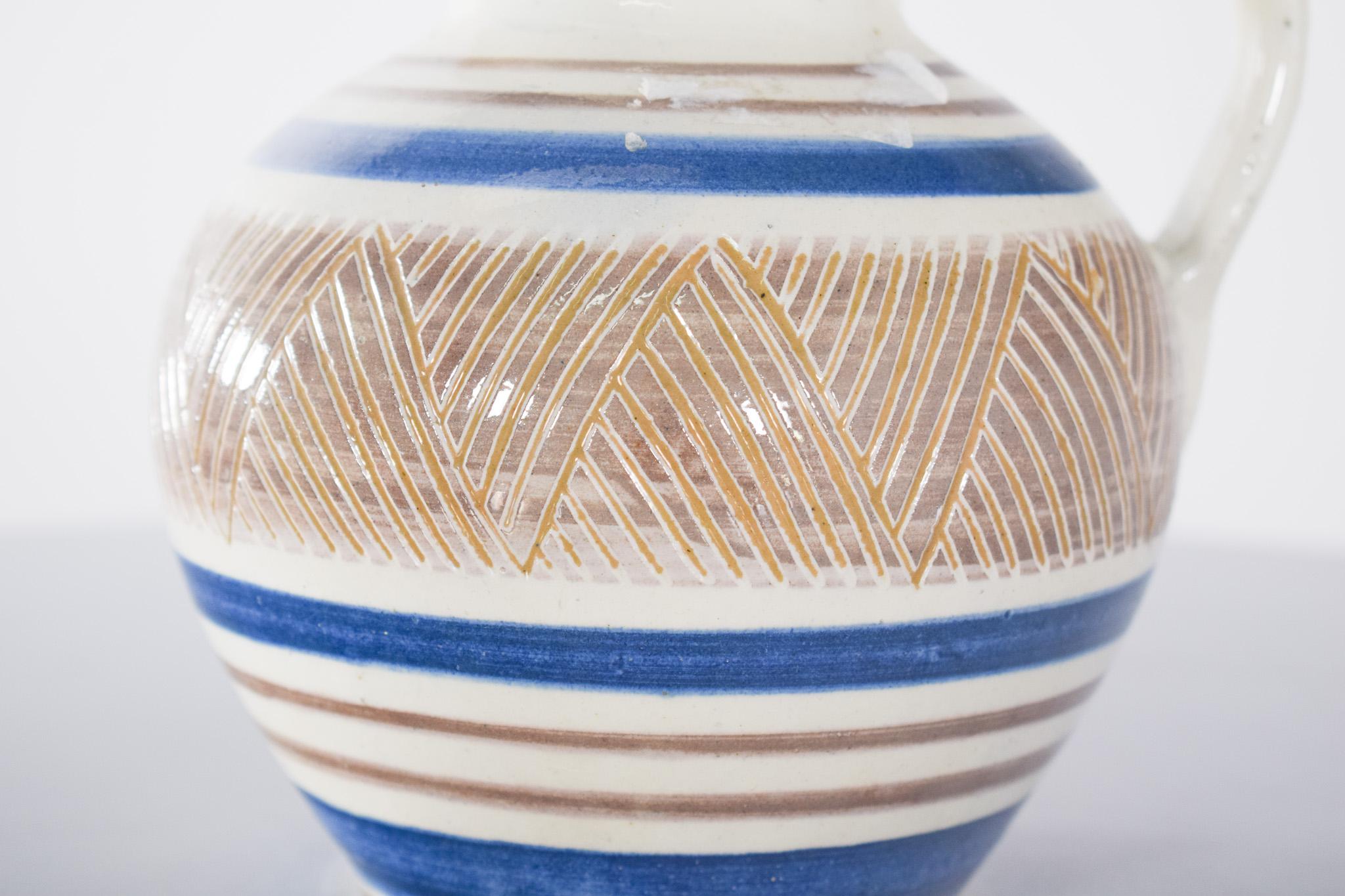 Brocca in ceramica a cupola. Presenta decorazioni tipiche del design scandinavo del XX secolo, che valorizzano la funzionalità unita alla sobrietà decorativa. Ceramica smaltata bianca, con strisce orizzontali nei toni del blu e della terra, la parte