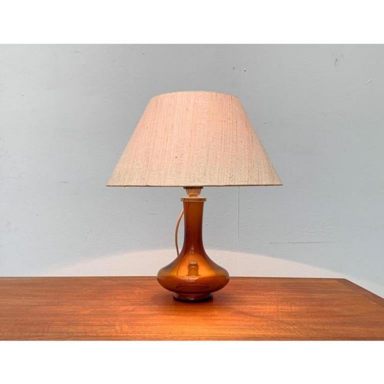 Rara lampada da tavolo danese del rinomato produttore Holmegaard.

Questa lampada da tavolo convince per la sua base ornamentale in vetro colorato. Il vetro trasparente è rifinito in colore ambrato. La trasparenza fa sì che la lampada corrisponda