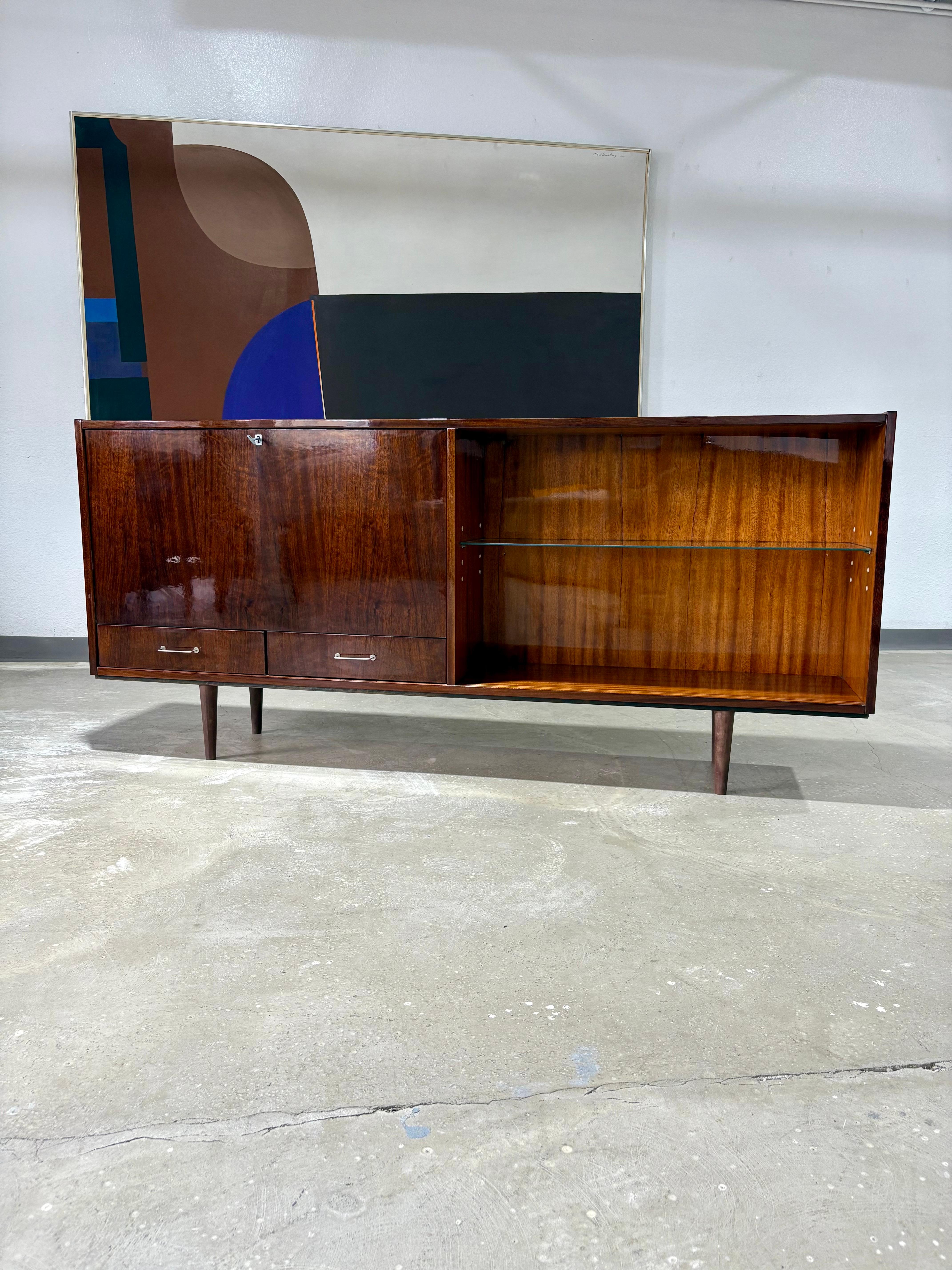 Credenza bar in mogano del Mid Century Modern Scandinavian. Presenta un'anta a ribalta con chiusura a sinistra e uno schienale interno diviso a specchio con un ripiano in vetro a sinistra e due ripiani in vetro regolabili a destra. Due cassetti con