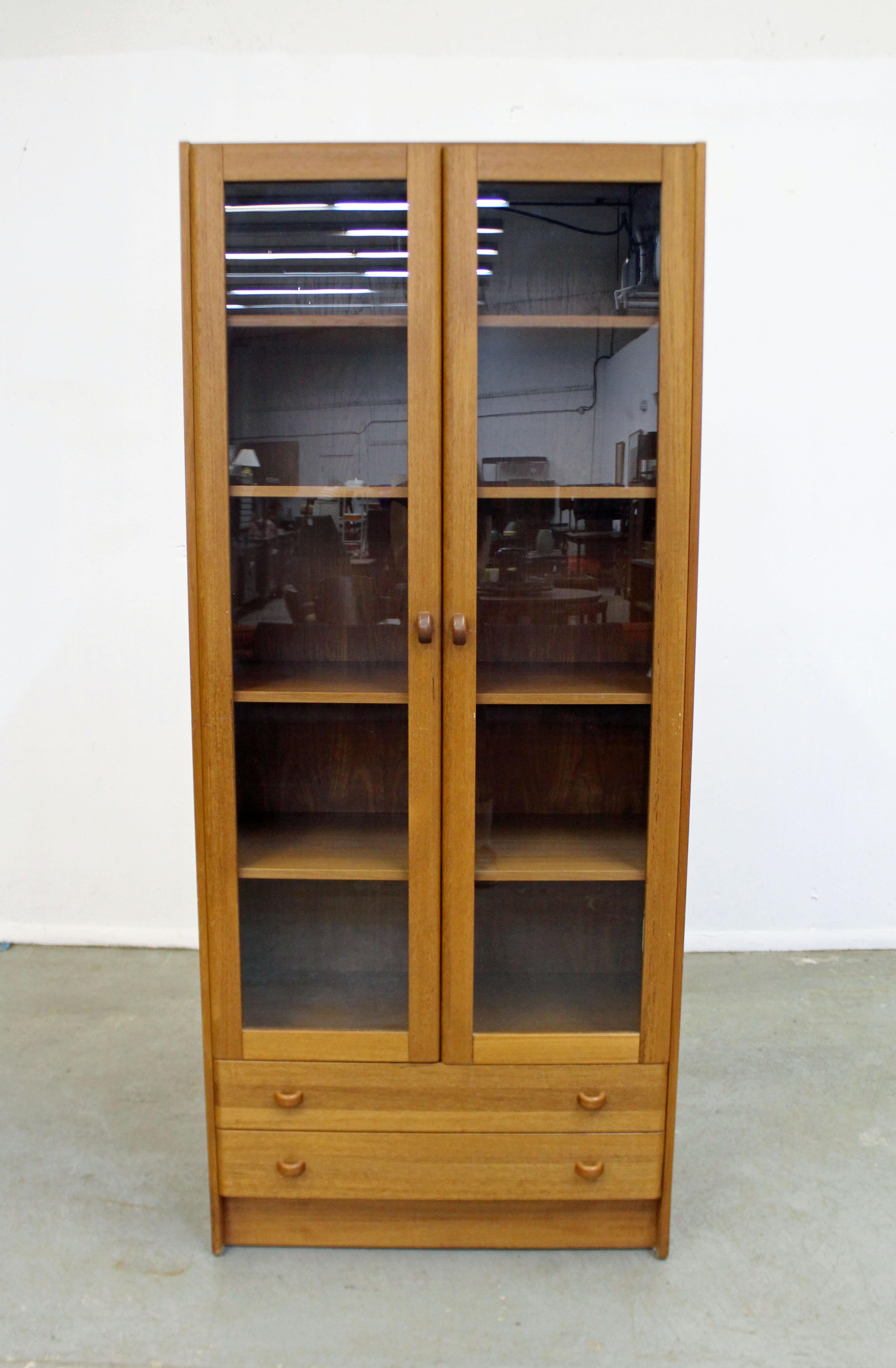 Offered is a Danish modern teak bookcase by Domino Mobler. This enclosed bookcase features two glass panel doors with three stationary shelves to the interior and two bottom drawers. It is marked on the back. See our other listings for more Danish