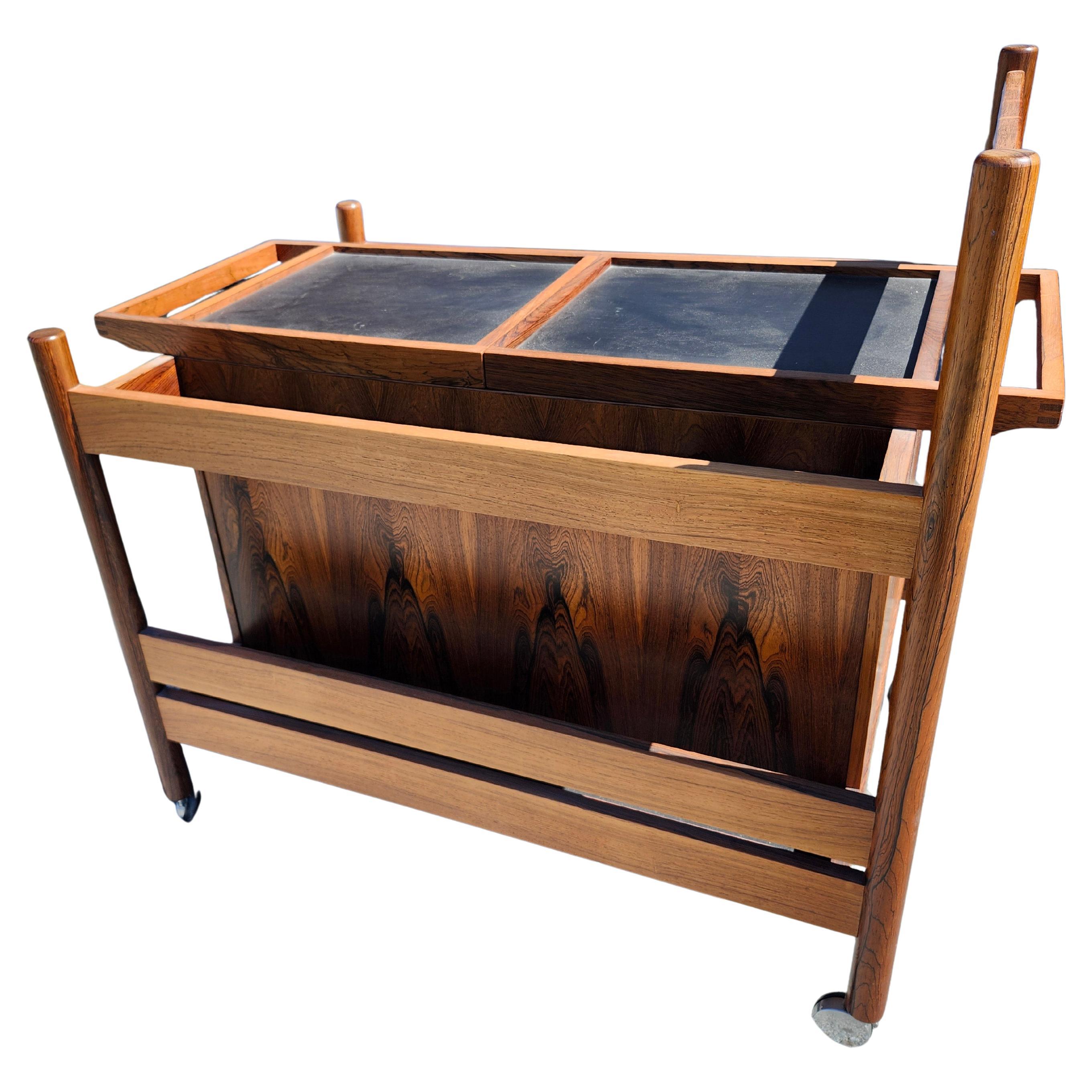 Fantástico carro de bar rodante con una zona de trabajo ajustable que se abre para revelar un amplio espacio de almacenamiento para toda tu cristalería y botellas. Bellamente diseñado en madera de palisandro  con encimeras laminadas para preparar