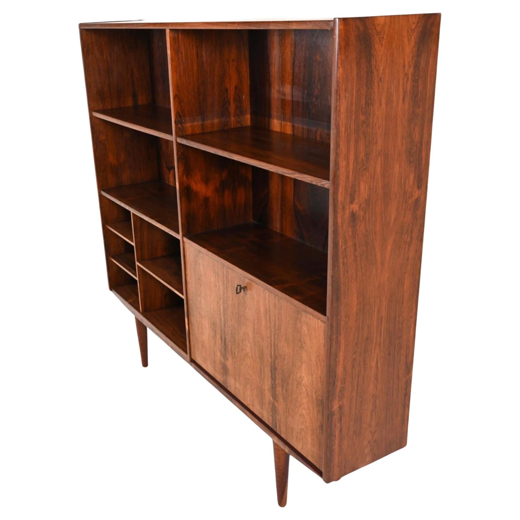Mid-century Rosewood wood wide and bookcase with 6 adjustable shelves and a lower right locking bar or desk storage area. Beautiful rosewood backing with Slim design and sits on 4 tapered teak legs. Made In Denmark. Has original key. Located in