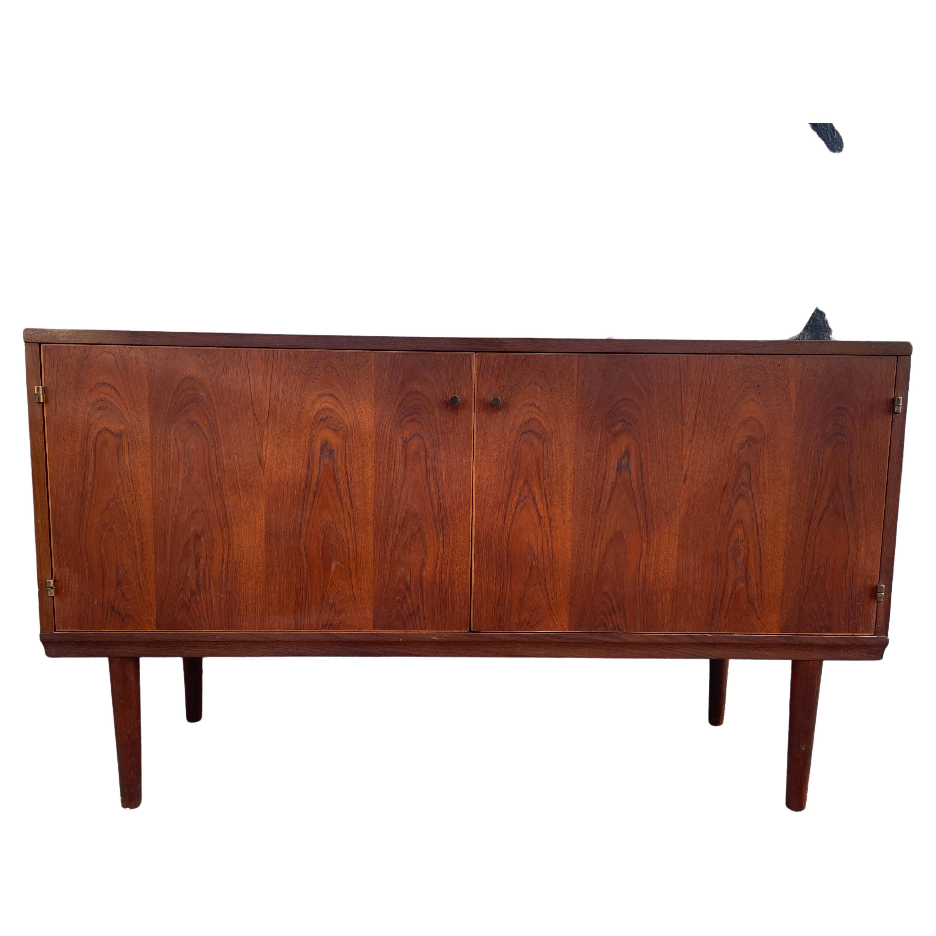 Rare Mid-Century Modern credenza finely crafted by LH mobler designed by Hans Olsen. All teak wood construction has (2) front cabinet doors with brass knobs and hinges. Has (2) adjustable shelves inside with (1) small drawer. Sits on (4) solid teak