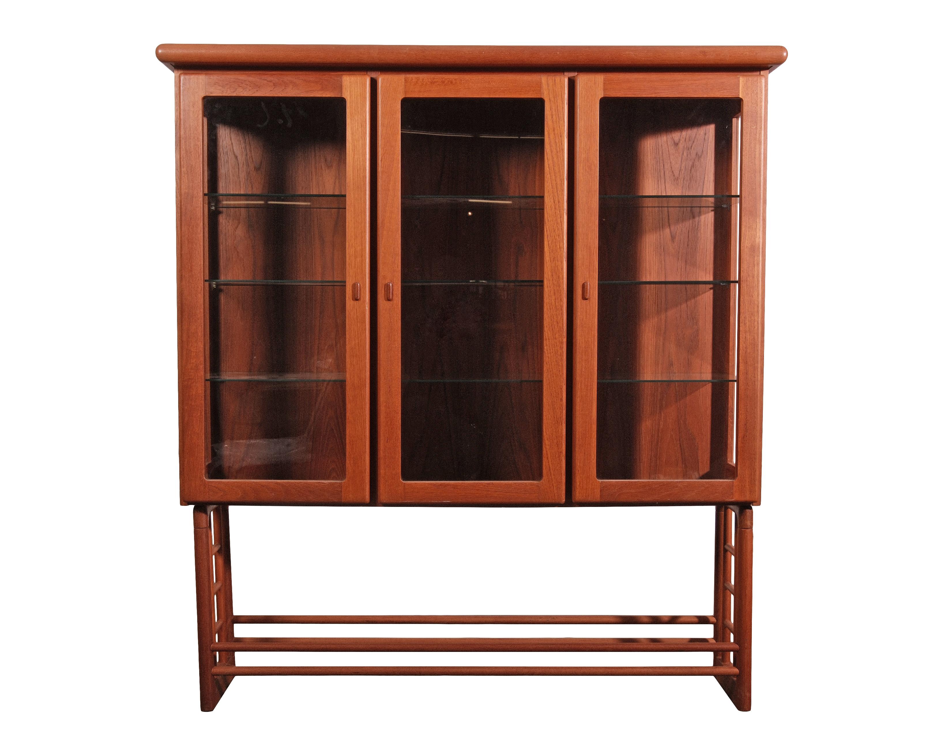 Questa raffinata vetrina in teak della modernità danese di metà secolo esemplifica l'eleganza e la maestria dell'epoca. La calda finitura in teak satinato completa l'elegante base con gambe a scaletta, mentre il triplo mobile a ripiani offre spazio