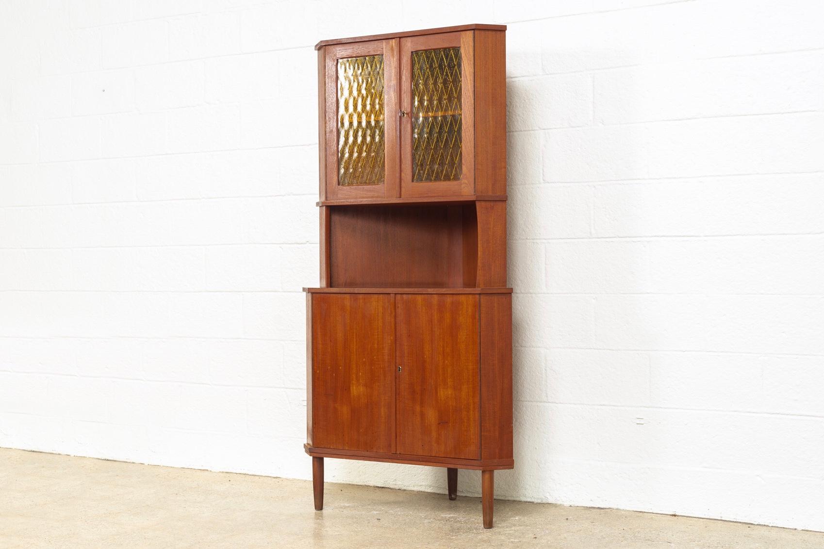 This vintage midcentury Danish modern teak corner cabinet is circa 1960. With classic Danish modern styling the cabinet has clean minimalist lines. This unique corner cabinet features a top hutch with beautiful leaded amber glass doors and one