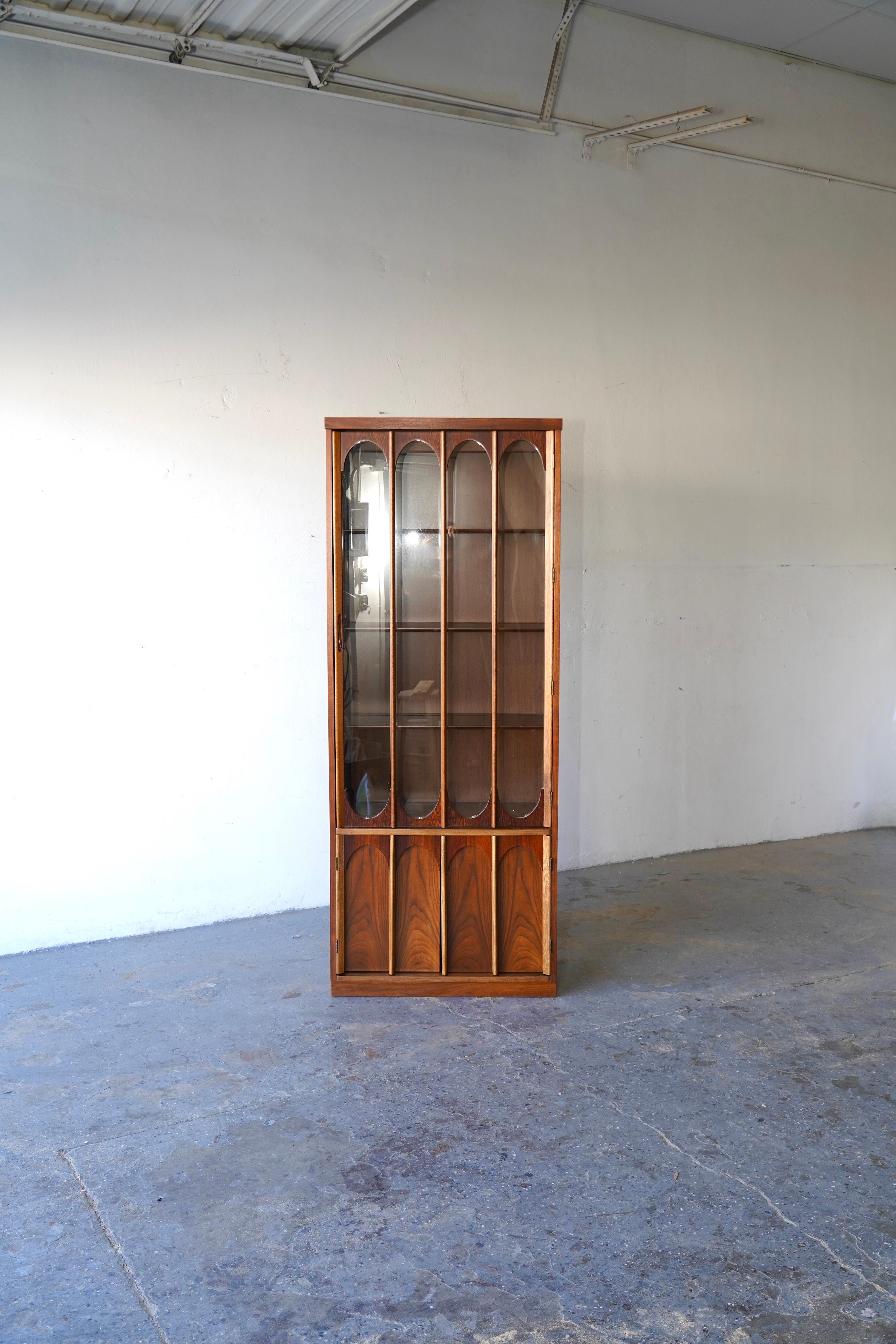This stunning Walnut & Rosewood  display cabinet, inspired by Broyhill design, showcases a distinctive sculpted aesthetic complemented by a spacious glass display with rosewood-accented arches. The cabinet also features enclosed storage below,