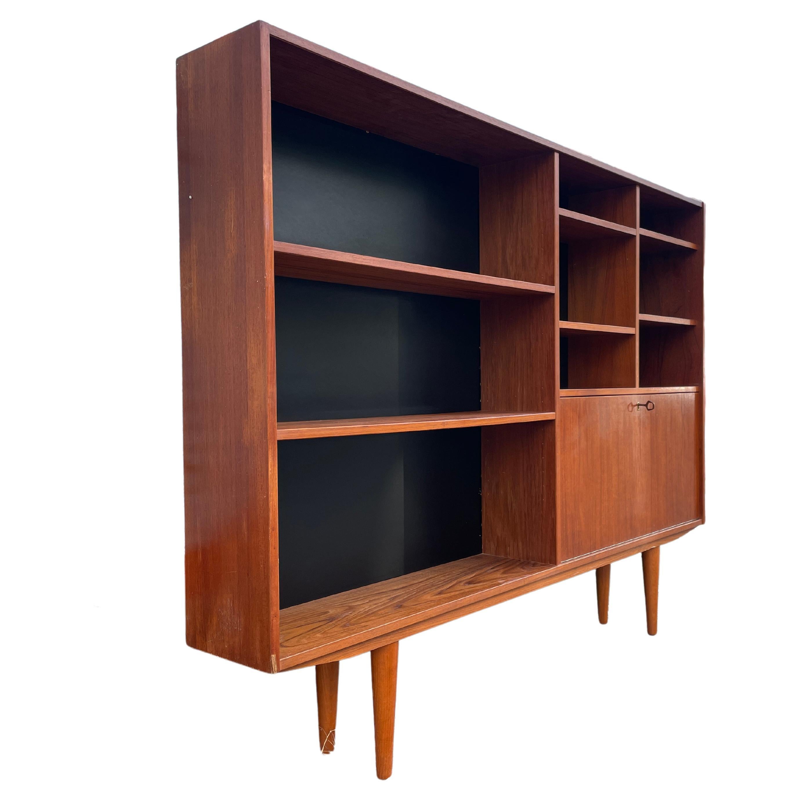 Mid-century teak wood wide and slim bookcase with 6 adjustable shelves and a lower right locking bar or desk storage area. Black backing with Slim design and sits on 4 tapered teak legs. Made In Denmark. Has original key. Located in Brooklyn