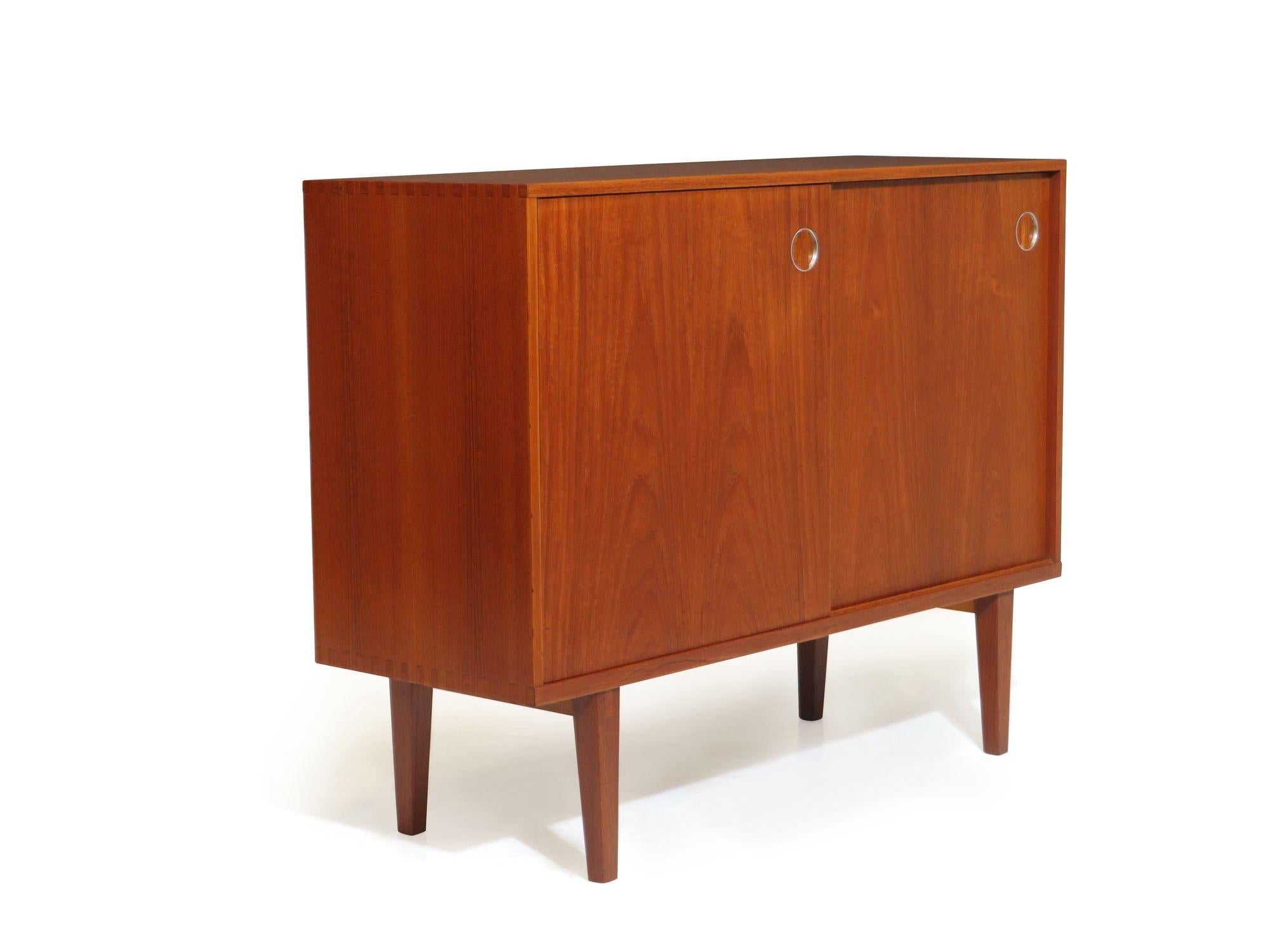 Mid Century Modern teak cabinet with pair of sliding doors with recessed oulls and metal details, adjustable interior shelves, and raised on tapered legs. Modest proportions make for a versatile cabinet appropriate as a night stand, media cabinet,