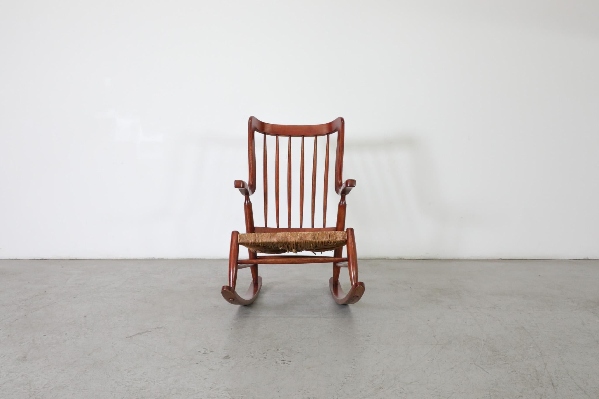 Mid-Century De Ster Gelderland Style rocking chair with spindle back, and rush seat. The frame is made from solid oak with a crimson stain and features sculpted organic curved armrests and frame. A blend of traditional style with classic materials