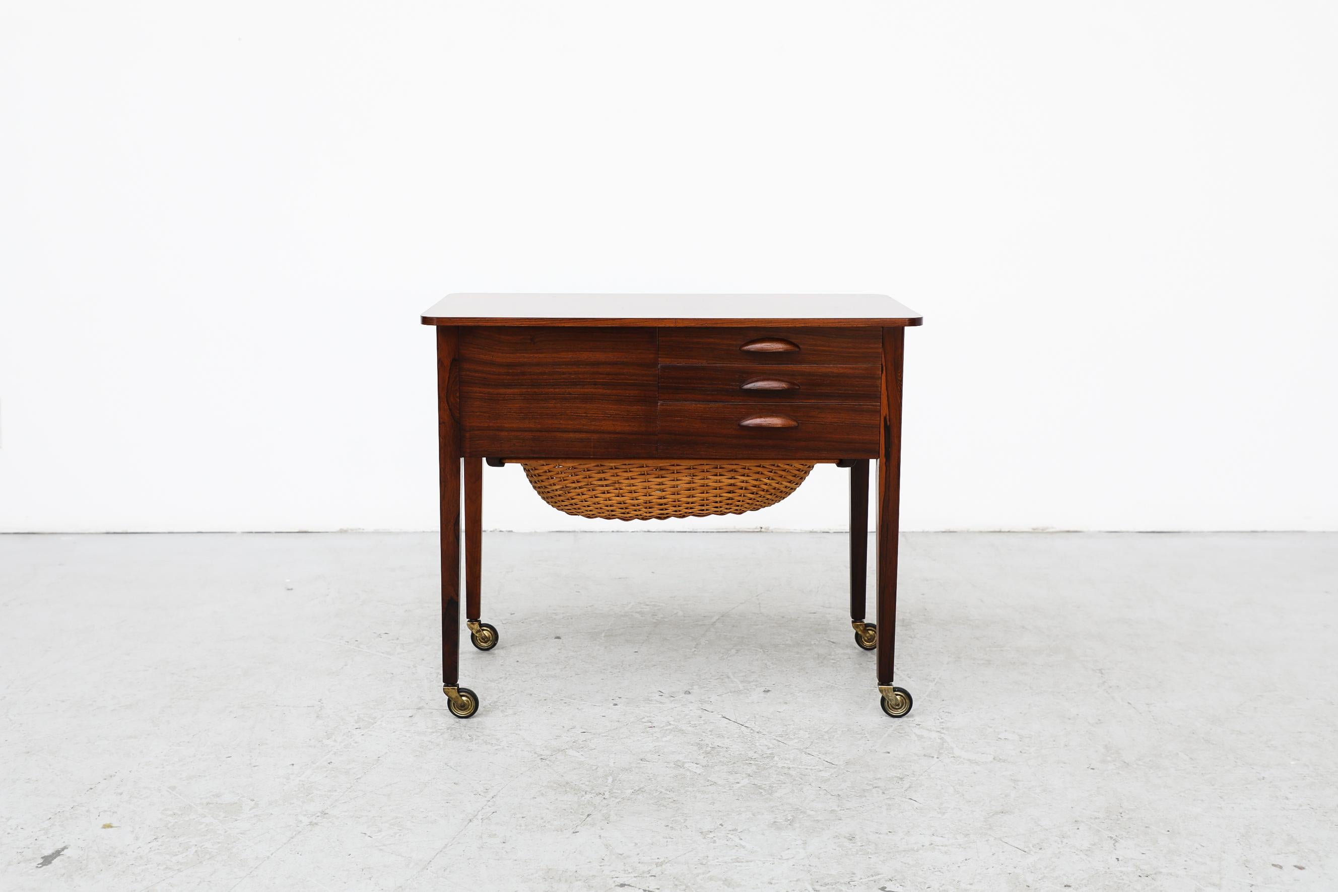 Charming Mid Century Danish Rosewood rolling side table with rattan sewing basket. The table has three front drawers, some with compartments and pegs for sewing gear and a bigger side drawer. In good original condition with normal wear consistent