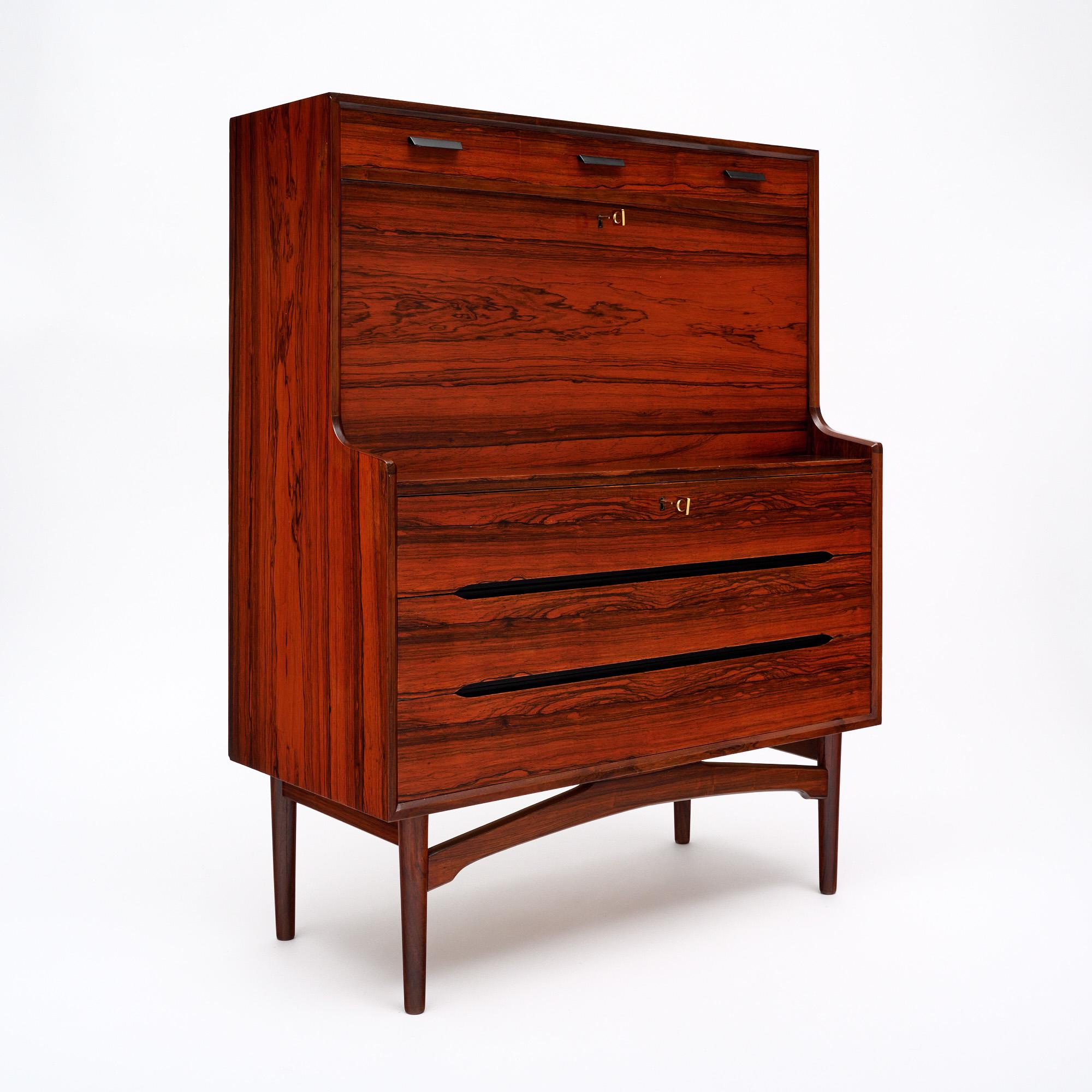 Secretario de Dinamarca de mediados de siglo, de construcción de madera maciza, principalmente palisandro. Esta pieza tiene cuatro patas de madera con una camilla en x. Hay tres cajones con cola de milano y tiradores ebonizados en la parte superior