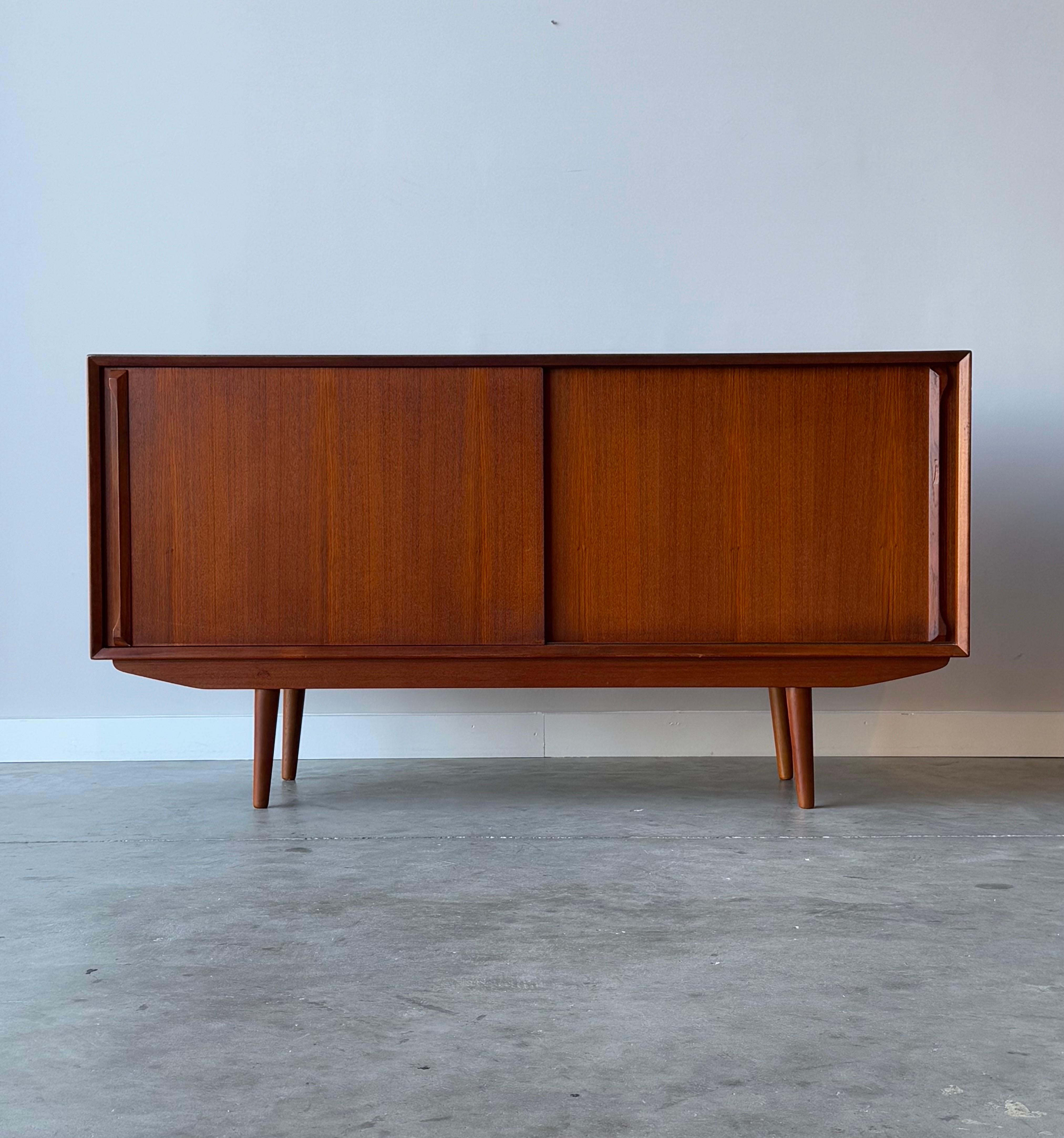 Mid-century danish credenza in the style of Arne Vodder. This case piece is made from teak wood and features two sleek sliding doors. Carved teak handles accent each door and allow for easy opening. The right-hand side opens to a long adjustable