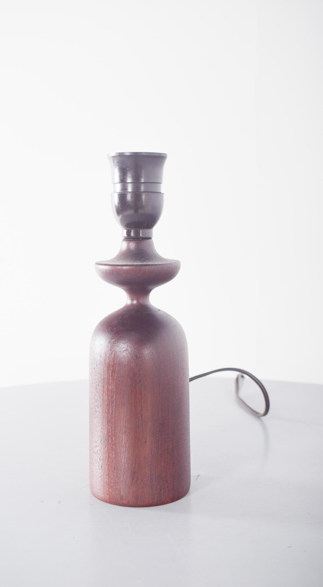 Lámpara de mesa, de madera de teca. La base tiene una forma cilíndrica redondeada que se estrecha hacia el centro, creando una silueta armoniosa. El acabado de madera barnizada refleja un aspecto cálido, realzando las vetas naturales de la teca. La