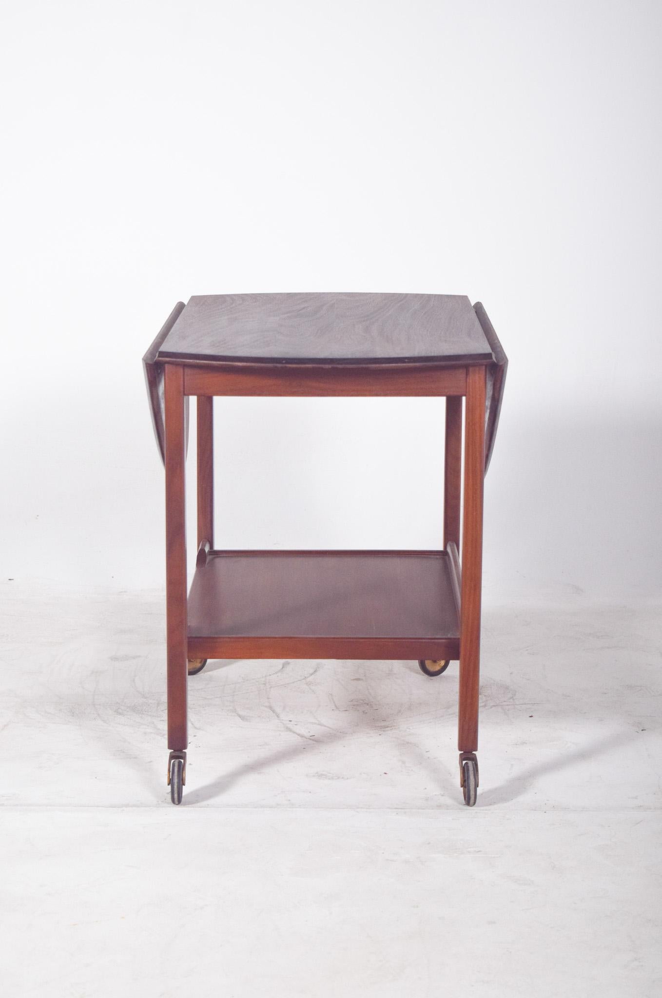 Tea trolley with flaps. Its slim, well-proportioned structure supports an oval top with hinged sides that can be raised to extend the support surface. The lower open shelf base extends the functionality of the piece without compromising the elegance