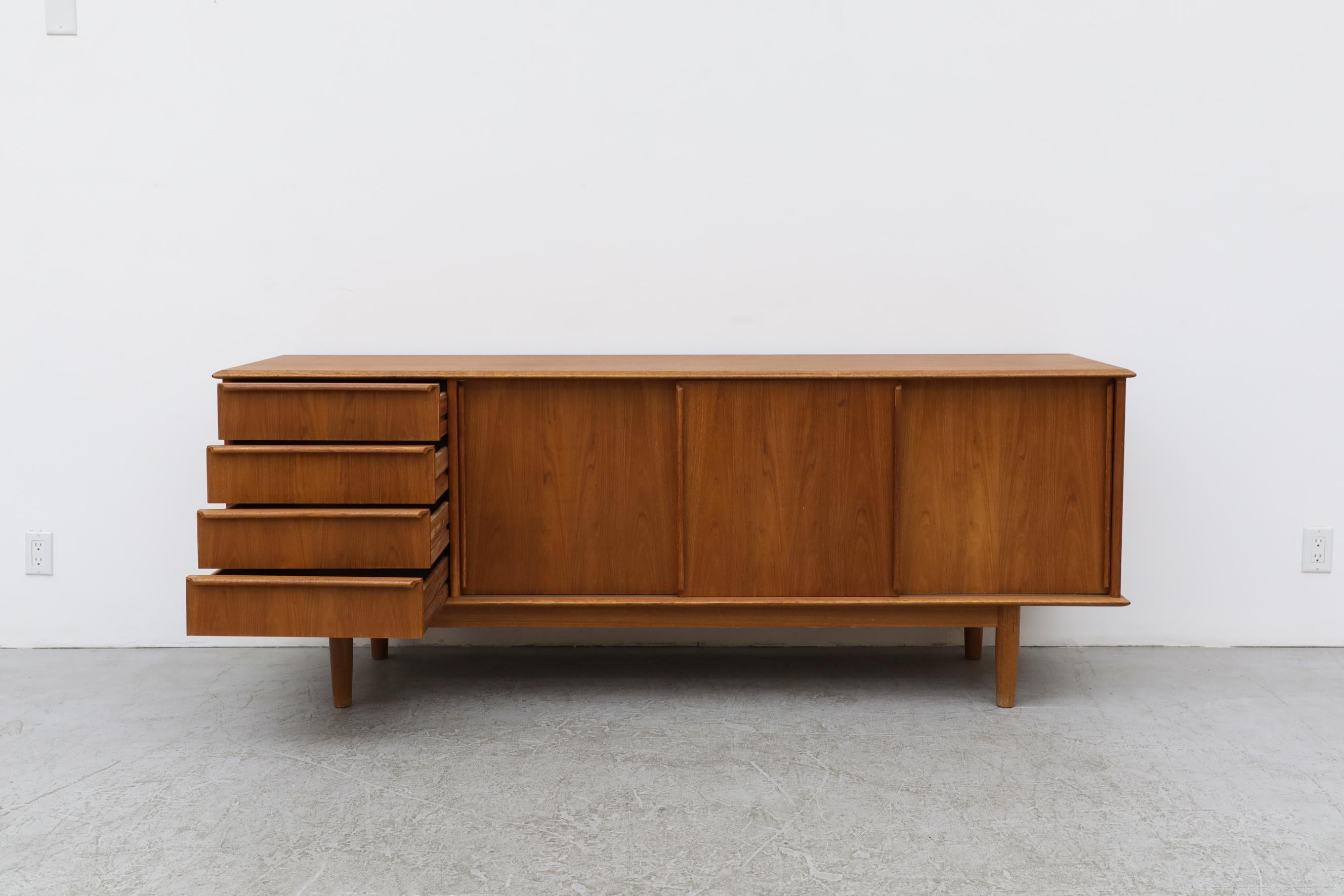 Long Danish teak credenza with long carved linear handles. The handles are vertical on the three sliding doors, and horizontally on the 3 side stacking drawers. In good original condition with visible wear including scratches and fading. Wear is