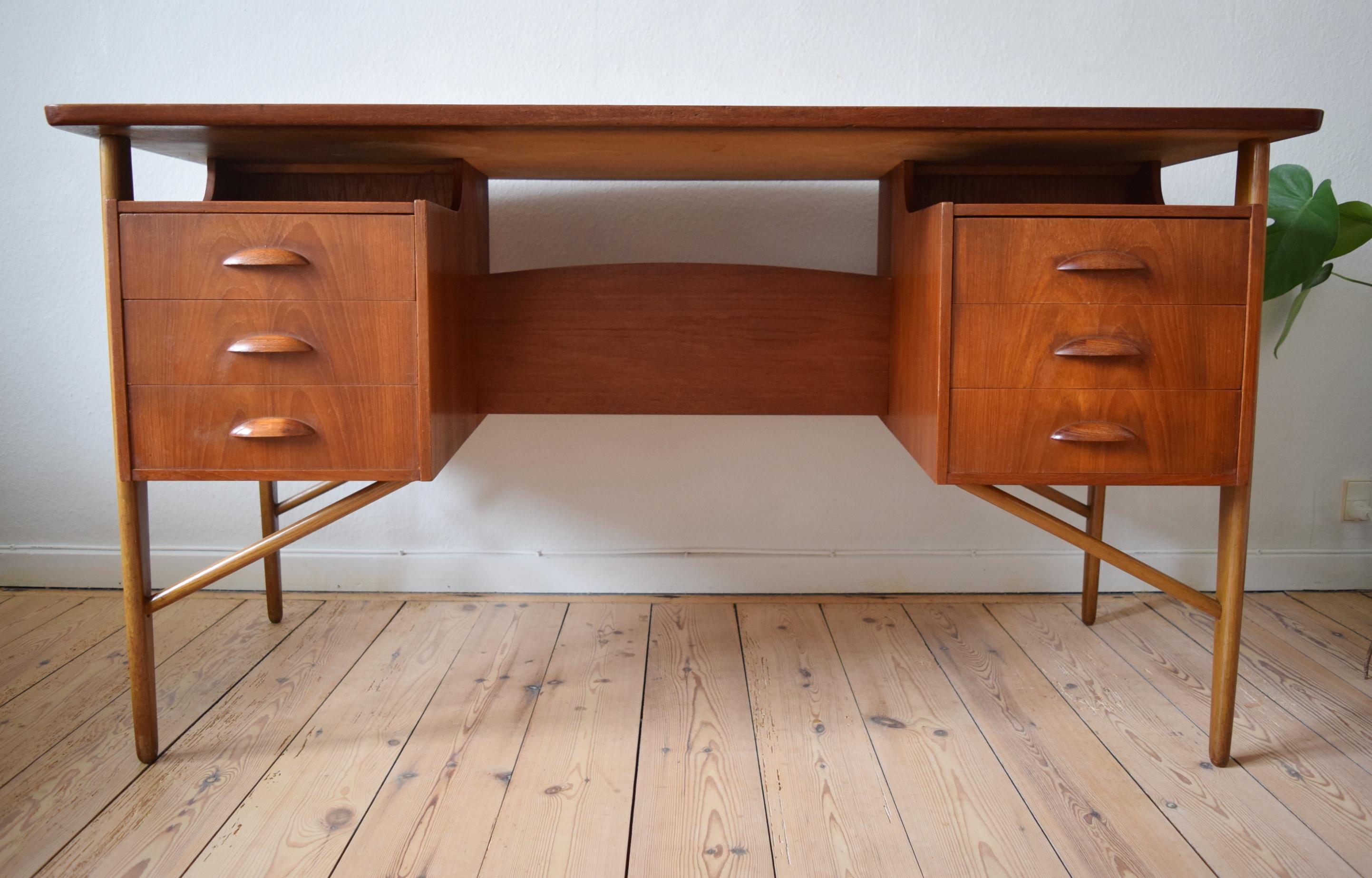 This teak desk was designed in the 1950s by Svend Aage Madsen for Sigurd Hansen. It features a curved top with a bevelled rear and side edges. Below there are six drawers with orange slice handles, and on the rear are two storage compartments and a