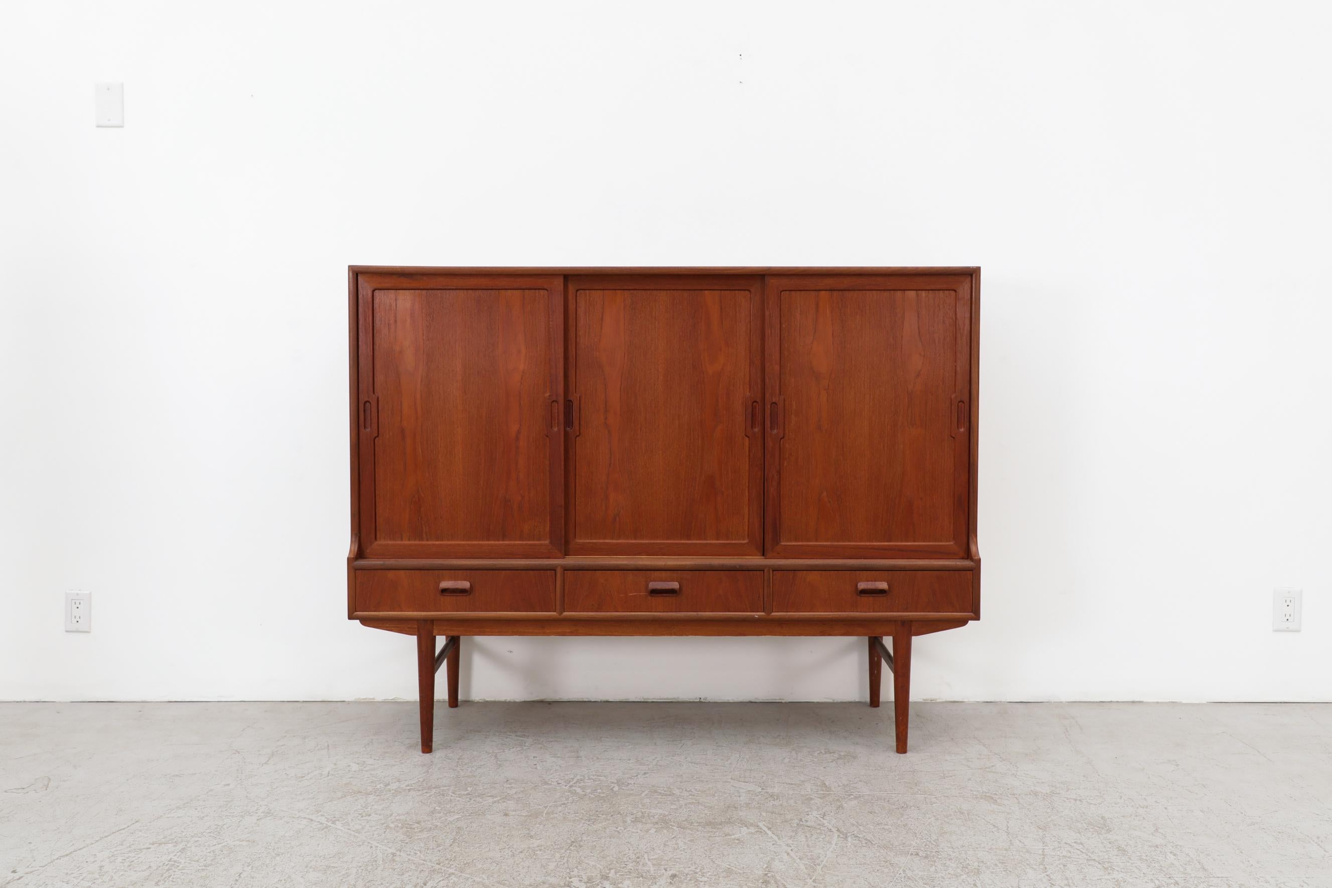 Danish Modern teak highboard with three sliding doors, inset carved oval pulls and plenty of storage space. Carved teak lower drawer handles, blonde interior wood, felt lined flatware drawers above with tapered legs. A lovely example of Danish