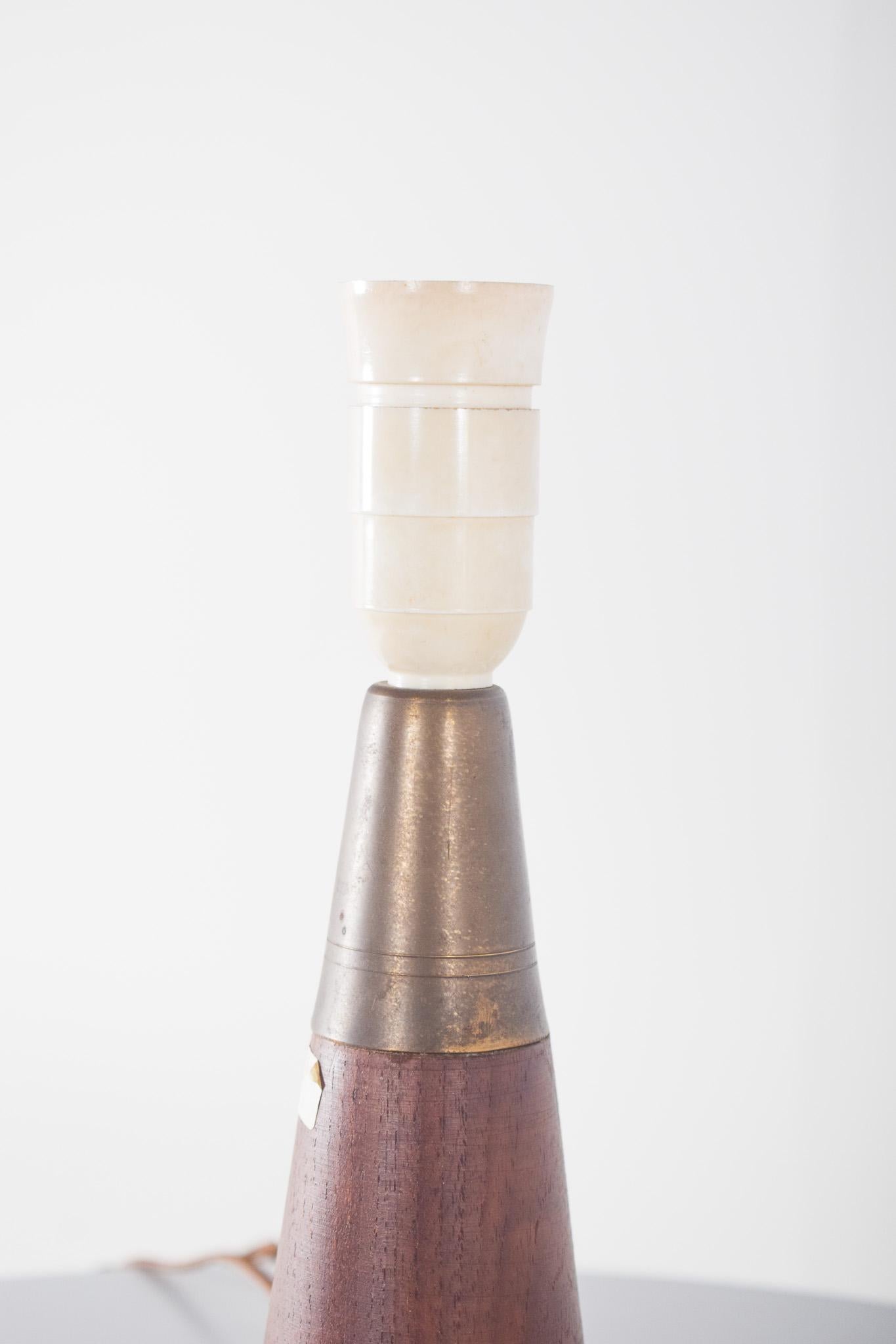 Table lamp, in teak wood. With the wood turned into a conical shape and finished in a warm tone, it highlights the natural grain of the wood. The structure is finished with a bronze-toned, slightly patinated metal fitting.
Two fine metallic lines