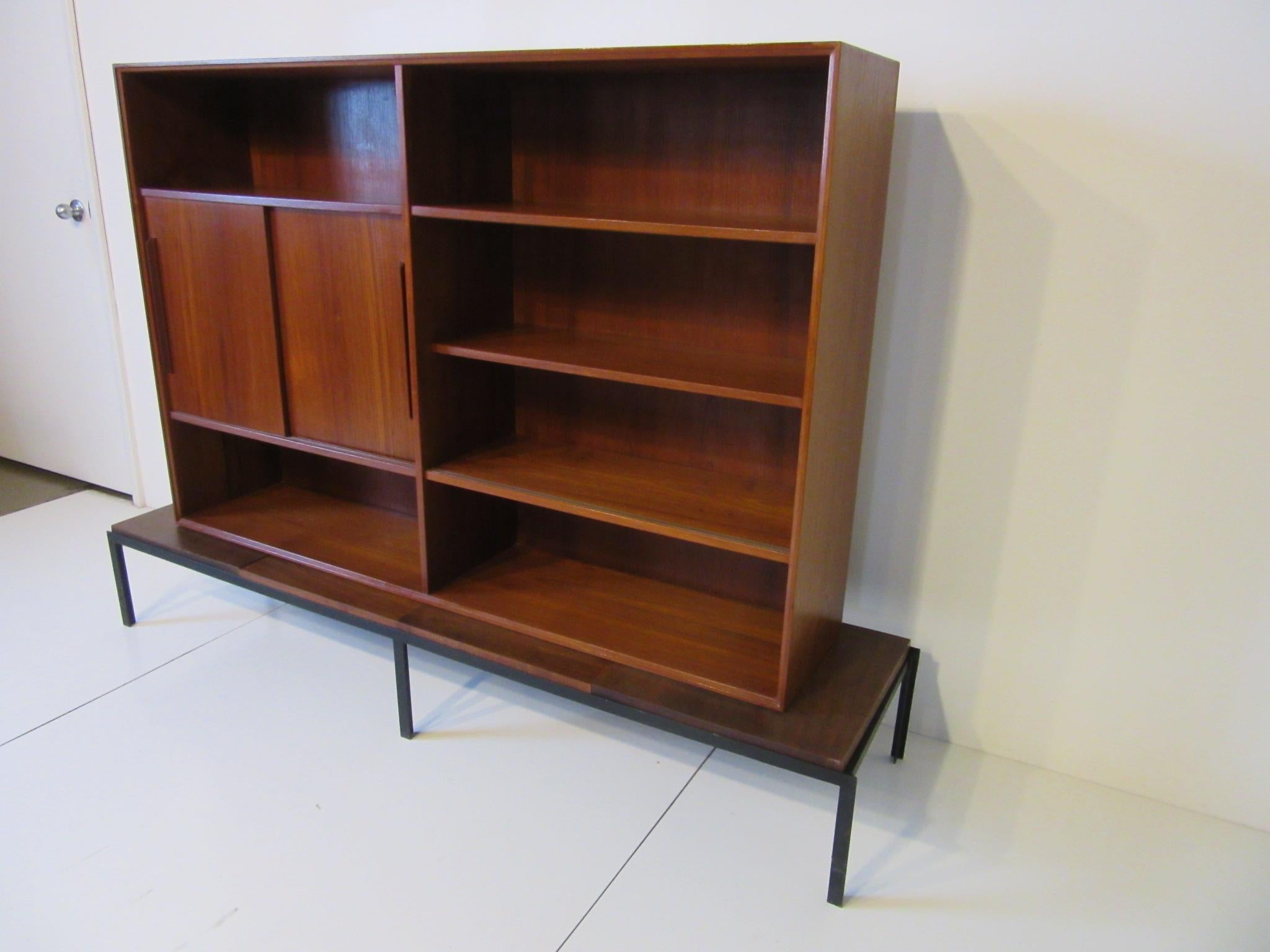 A two-piece midcentury bookcase with upper teak section, four adjustable shelves and two non adjustable shelves behind double sliding doors. The lower bench styled piece has four drop in walnut top panels sitting on a satin black metal frame. The