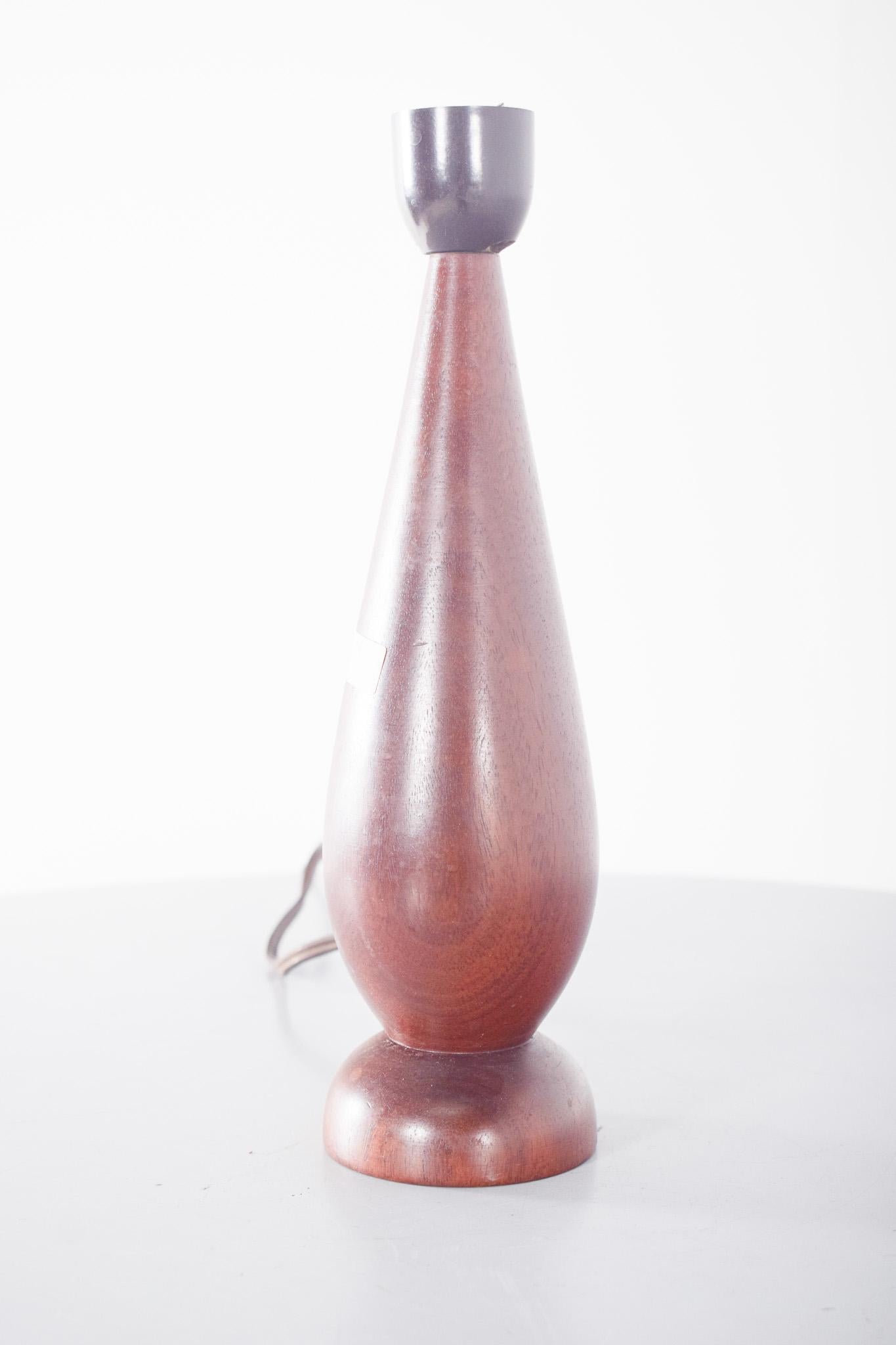 Lampe de table en bois de teck. Base en bois massif tourné, aux lignes élégantes et à la forme conique inversée, s'élargissant au niveau de la base arrondie. Le bois a une finition polie et vernie, avec une tonalité chaude, mettant en valeur le