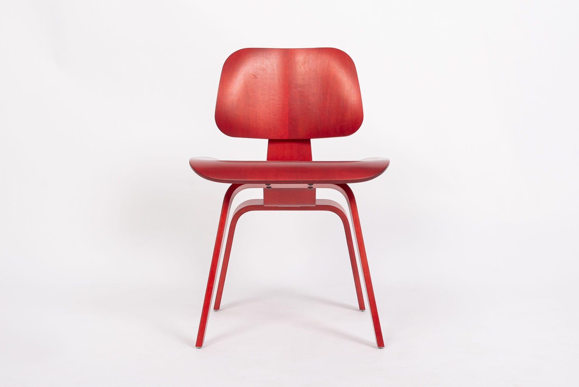 This authentic molded plywood DCW (Dining Chair Wood Base) dining chair by Charles and Ray Eames for Herman Miller is expertly crafted from red-stained ash molded plywood and features a molded five-ply seat and back, eight-ply legs and back brace
