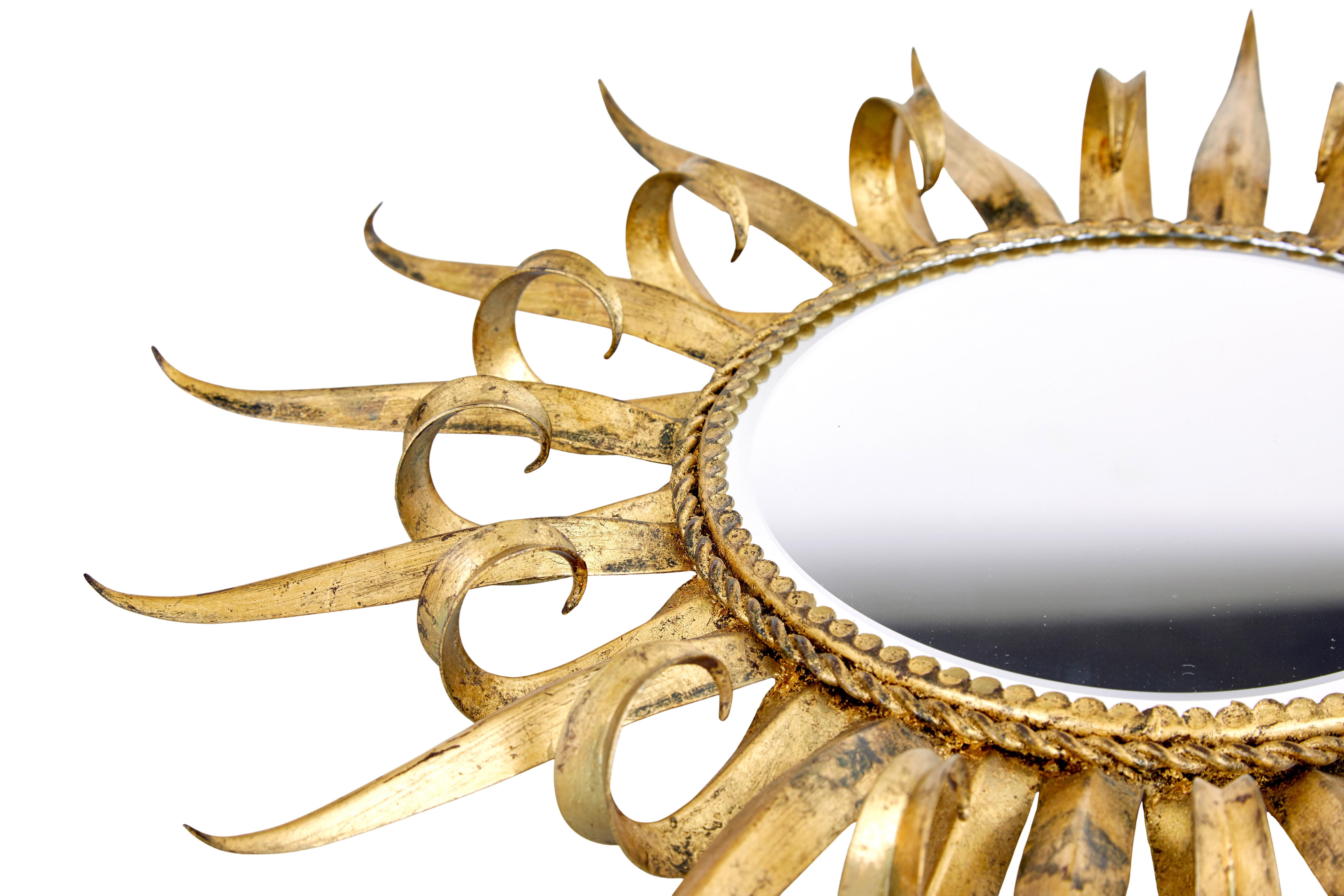 Mid century decorative sunburst mirror circa 1970.

Scandinavian deco revival wall mirror.  Circular mirror with bevelled edged, beaded metal border, surrounded by alternating straight and curling metal leaves.  Presented in a weathered gold effect