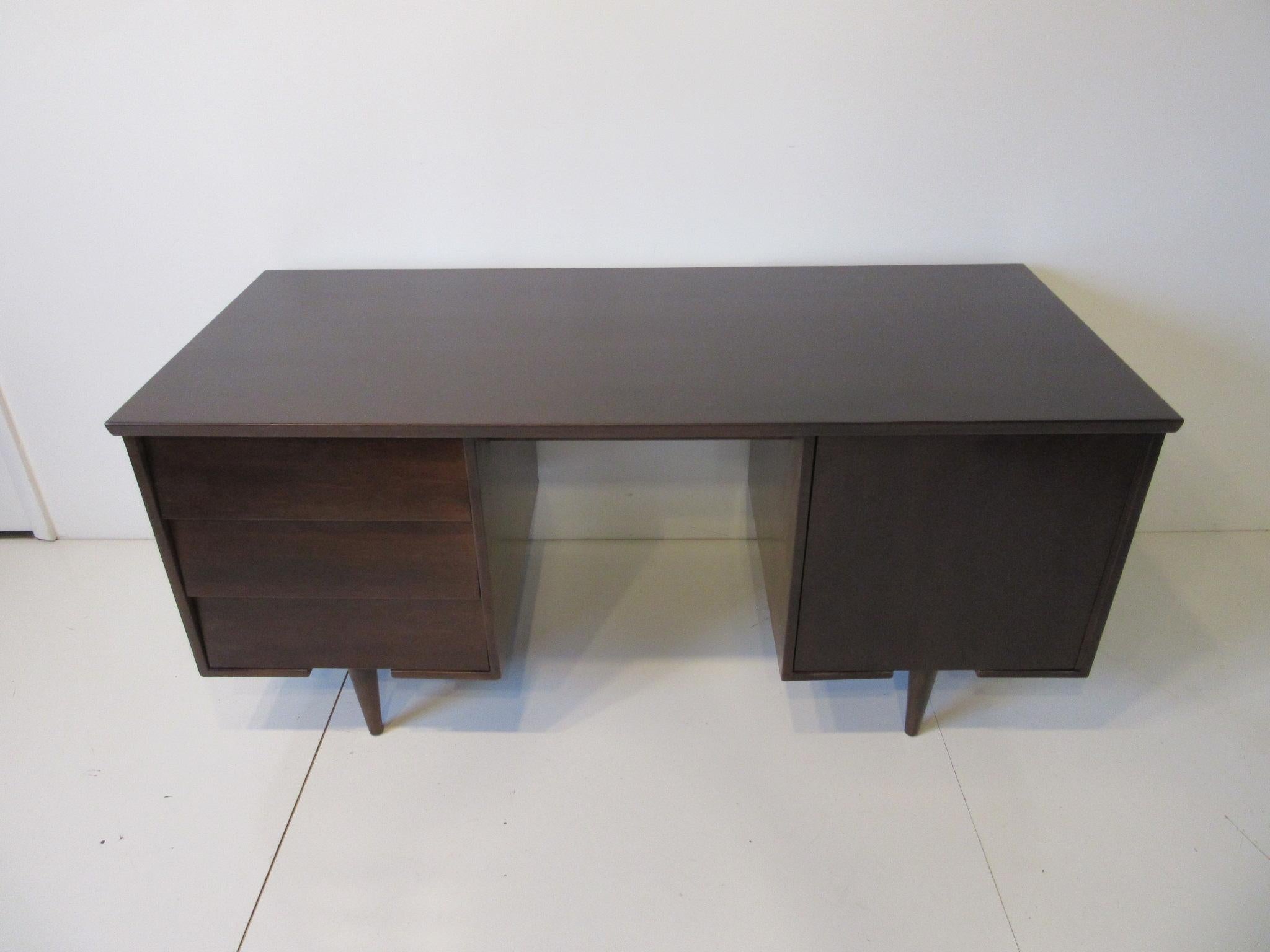 A dark ebony toned wood midcentury desk with three drawers the top one having pen/ pencil divider. The other side has a door and flat file storage with one removable shelve, retains the manufactures ink stamp to the drawer bottom Mengel Bord