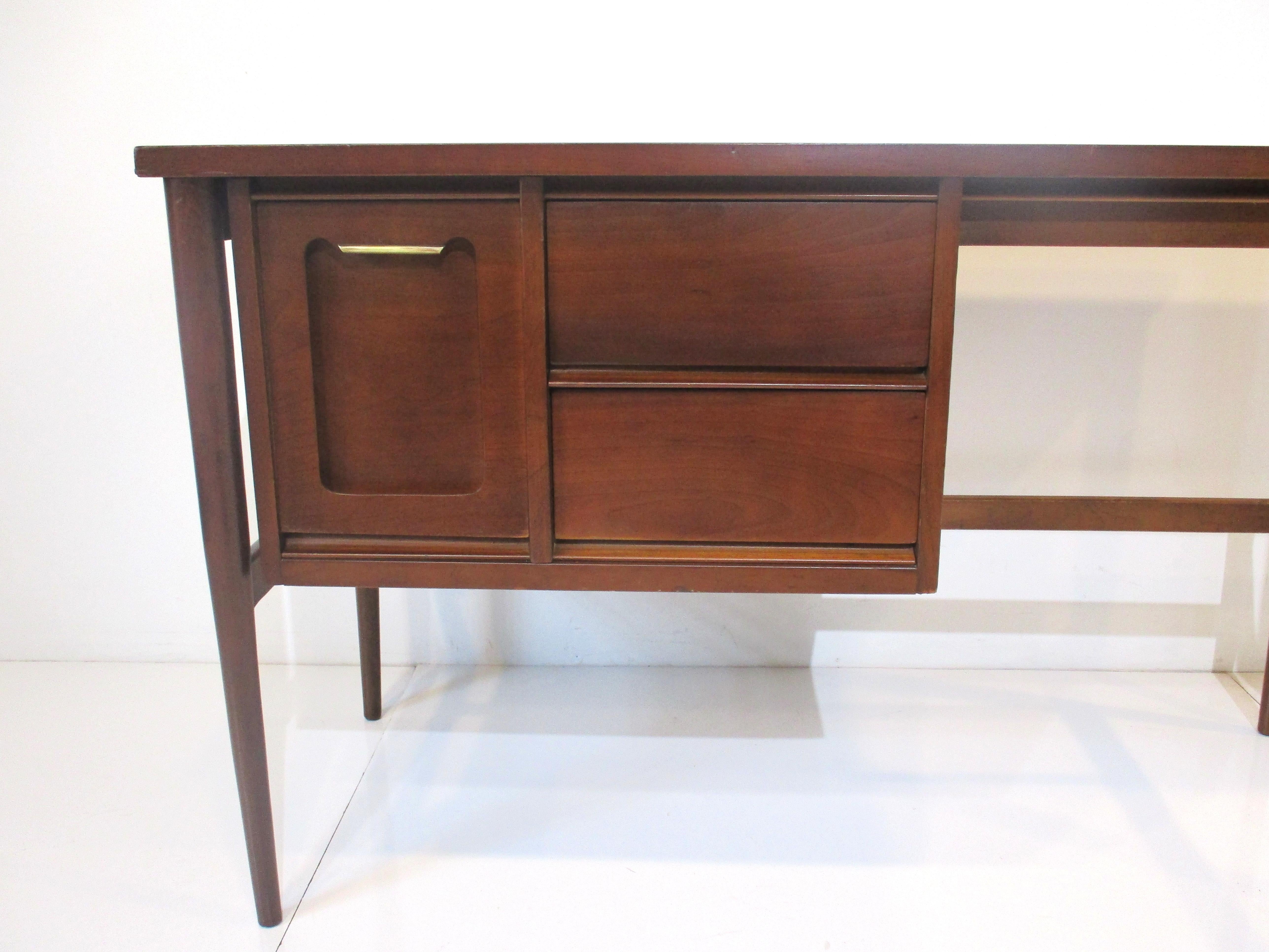 A medium walnut toned mid-century desk with a file drawer having a cutout design detail to the front and a thin upper brass pull and two regular drawers . The kneehole has a lower cross stretcher and conical legs manufactured by the Bassett