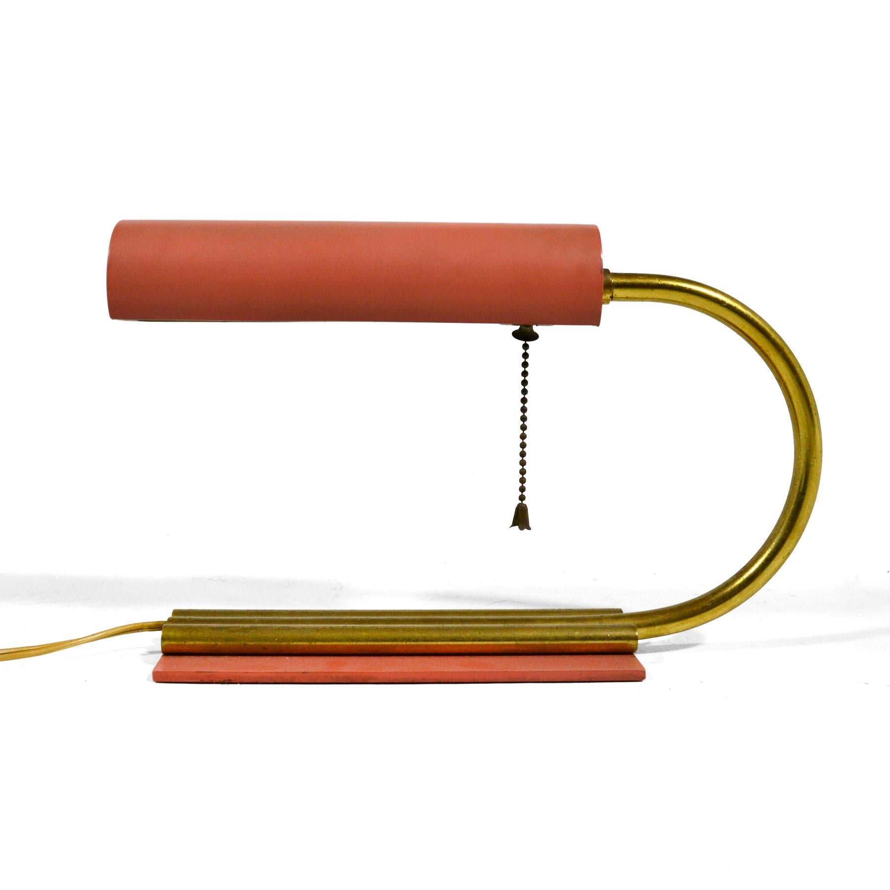 This elegant little desk lamp has an early midcentury or streamline moderne aesthetic in the manner of Gilbert Rohde. The C-shaped curved brass neck connects the rotating cylindrical head to the base and provides a cord chase. The wonderful salmon