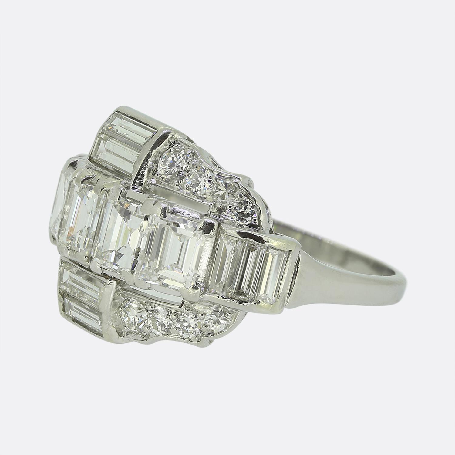 Aquí tenemos un notable anillo de racimo tomado a mediados del siglo XX. Esta pieza de platino luce cinco brillantes diamantes blancos talla carre en el centro de la esfera. Estas piedras principales se complementan con un dúo de diamantes baguette