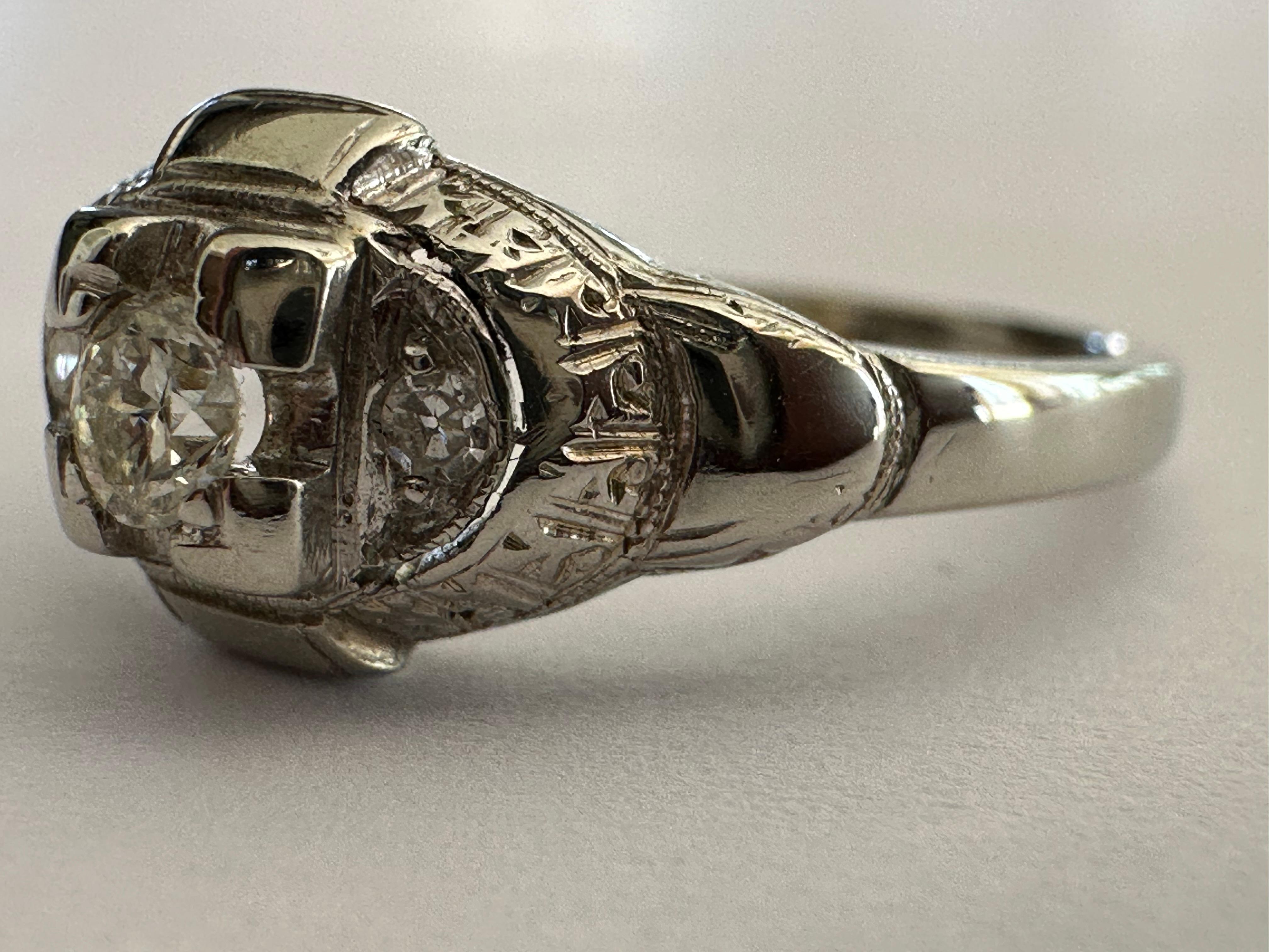 Une pierre centrale en diamant de taille européenne ancienne mesurant environ 0,10 carat, de couleur I et de pureté VS, brille dans une monture carrée rehaussée de deux diamants de taille unique et d'une délicate gravure à la main. Serti en or blanc