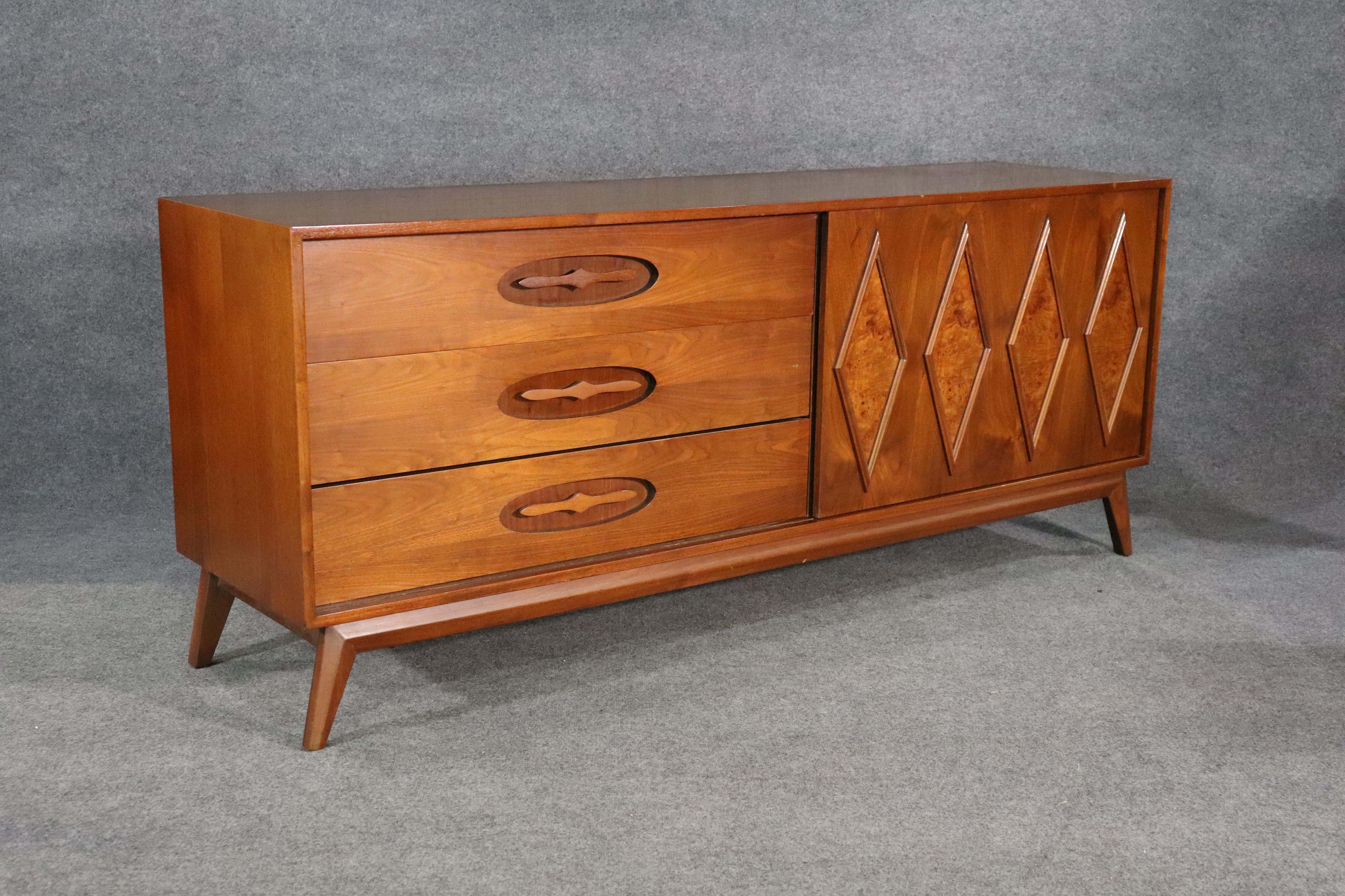 Long American made dresser with sliding door. Six wide drawers, sculpted inset handles and burl veneer diamond shaped accents.
Please confirm location NY or NJ