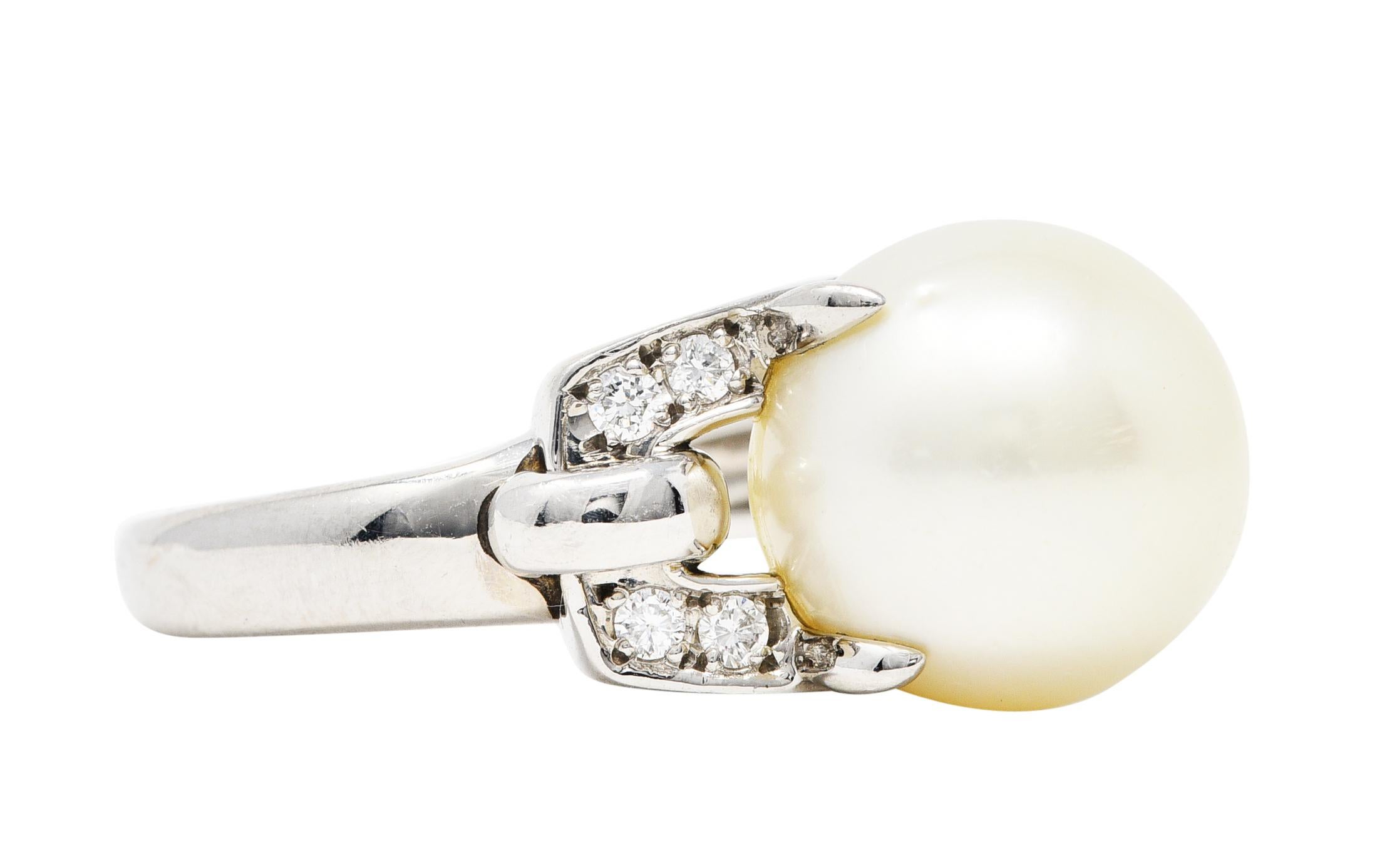 Cocktail style ring designed with high polish and buckle motif

Centering a prong set 12.0 mm round pearl - white in body color with good luster

Flanked by buckle motif shoulders each accented by four round cut diamonds

Diamonds weigh