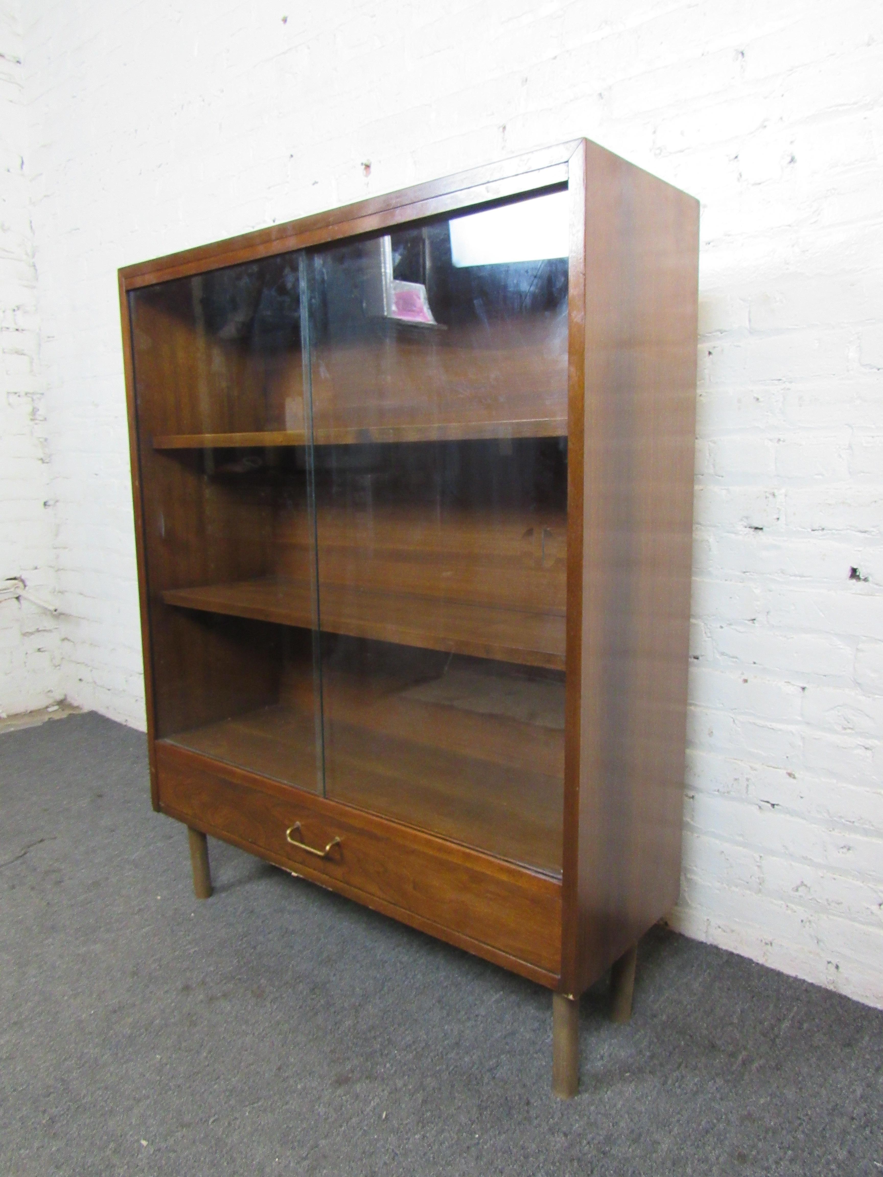 This large cabinet by Martinsville can be used as a display case or for storage with its large amount of shelf space and sliding glass doors. Handsome walnut woodgrain elevates this cabinet's design, and a large bottom drawer allows for further